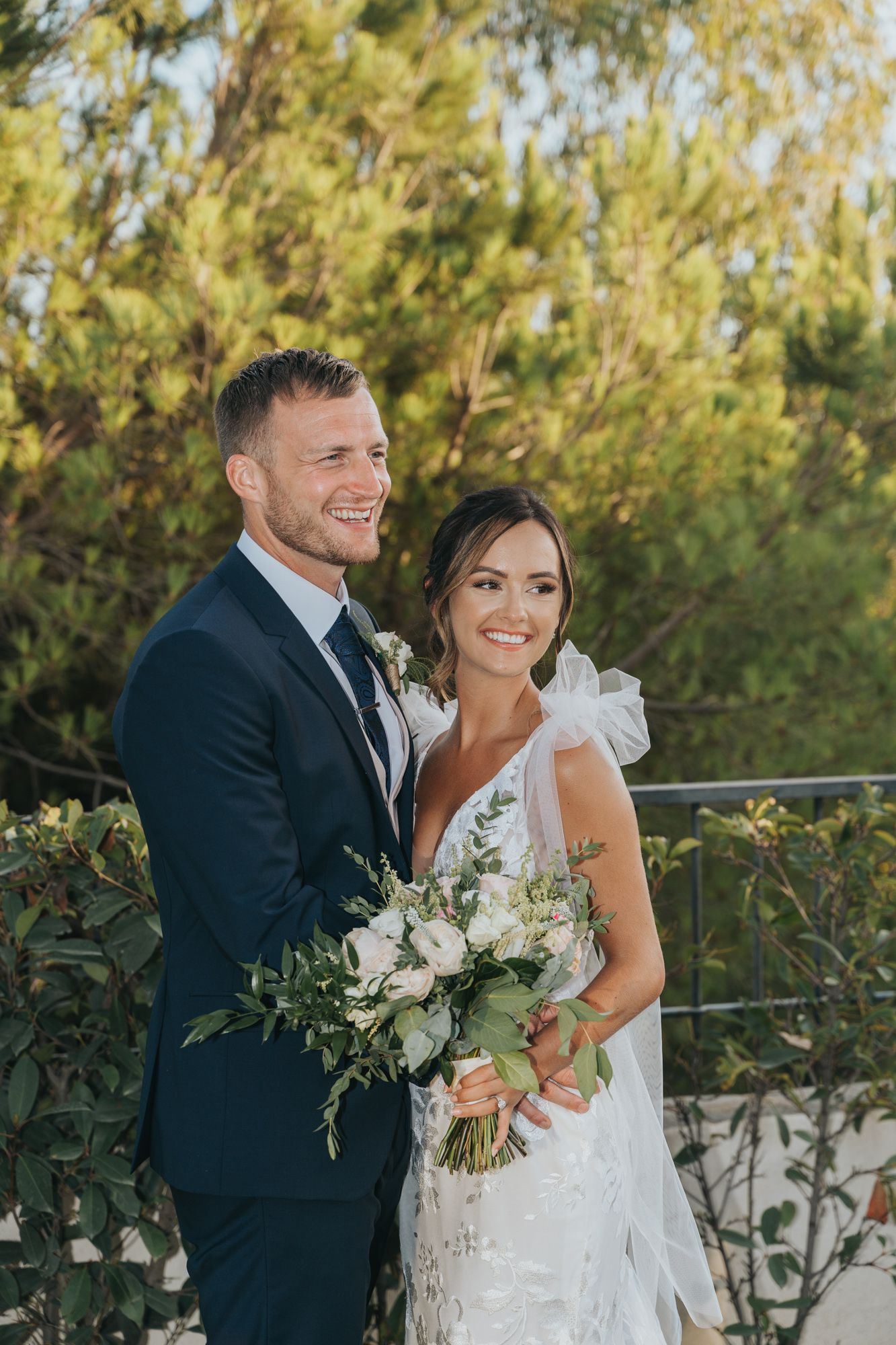 A couple poses together outdoors on their wedding day with a white floral bouquet and greenery in the background.