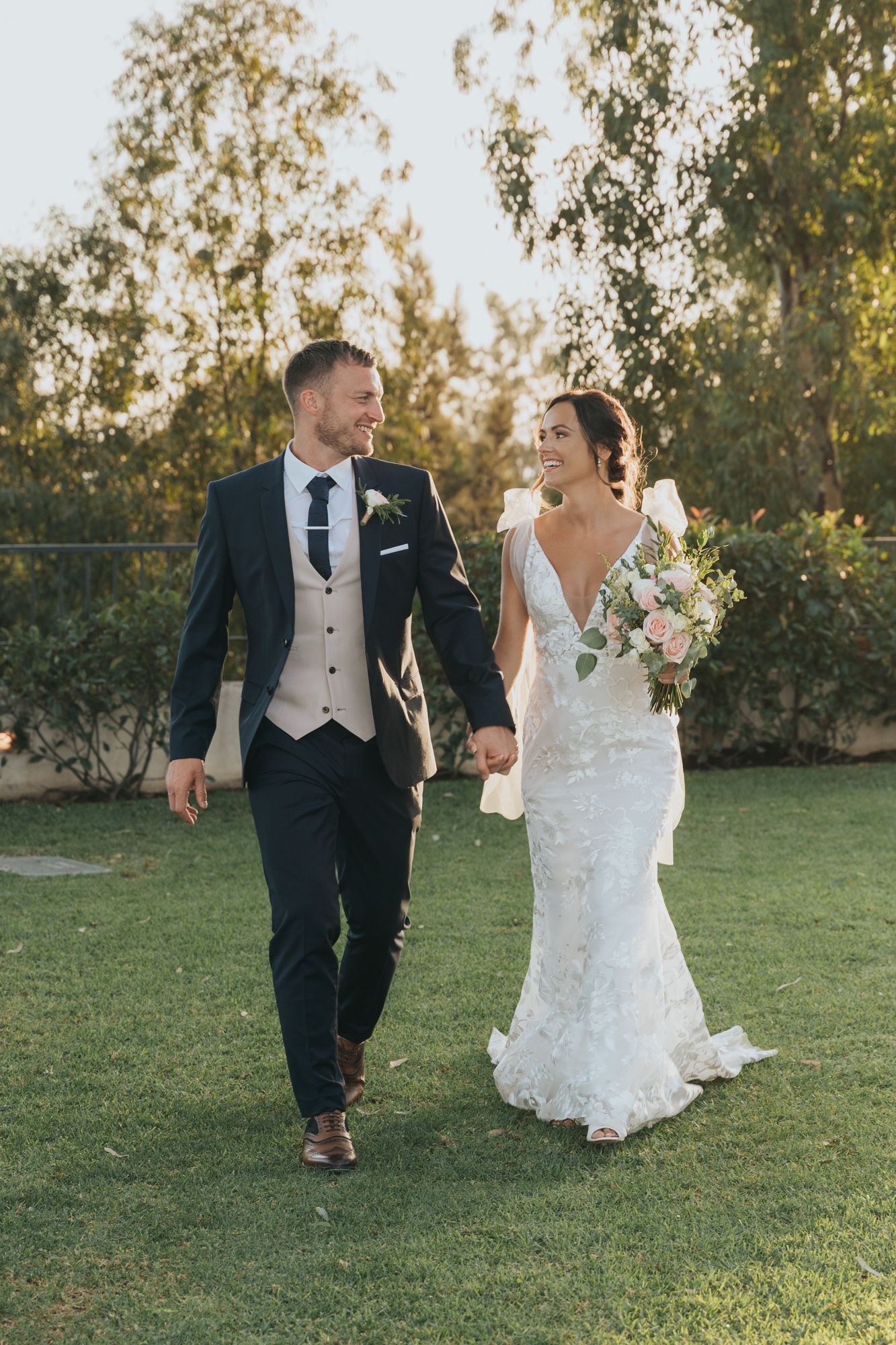 A newlywed couple walks hand in hand on a grassy lawn during golden hour, the bride holding a soft pink bouquet.