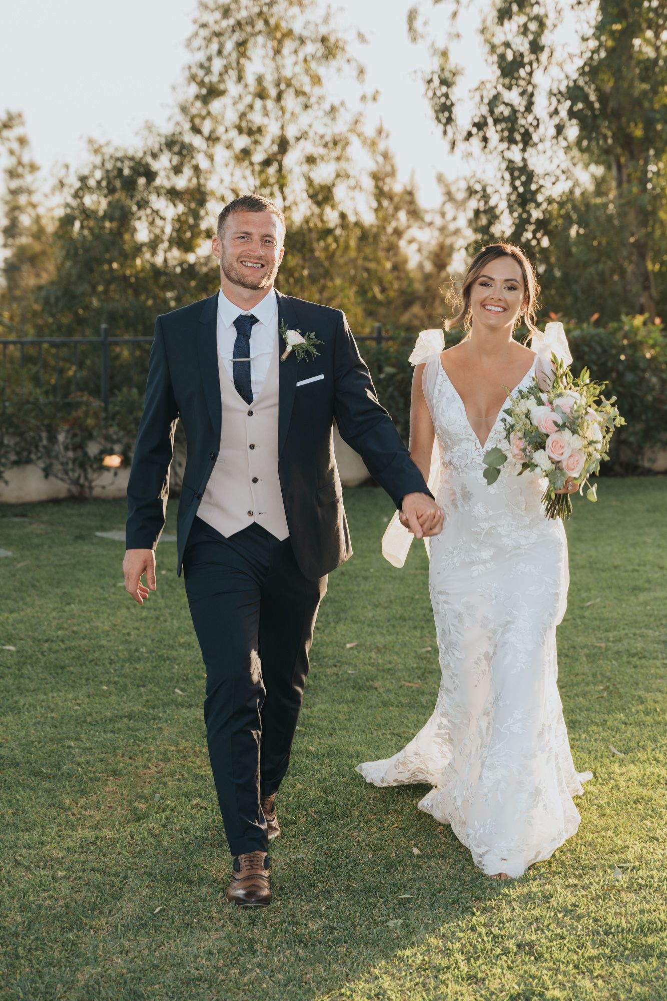 A bride and groom walk together on grass, with the bride holding a pink and white floral bouquet.