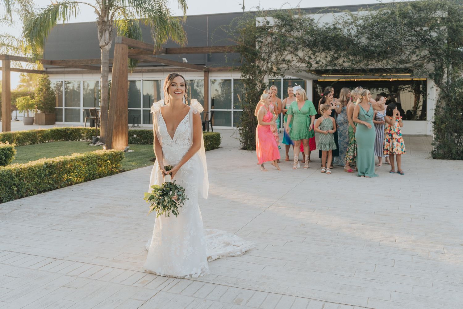 A bride in a white wedding dress holds a bouquet while bridesmaids in coral and mint dresses stand in the background.