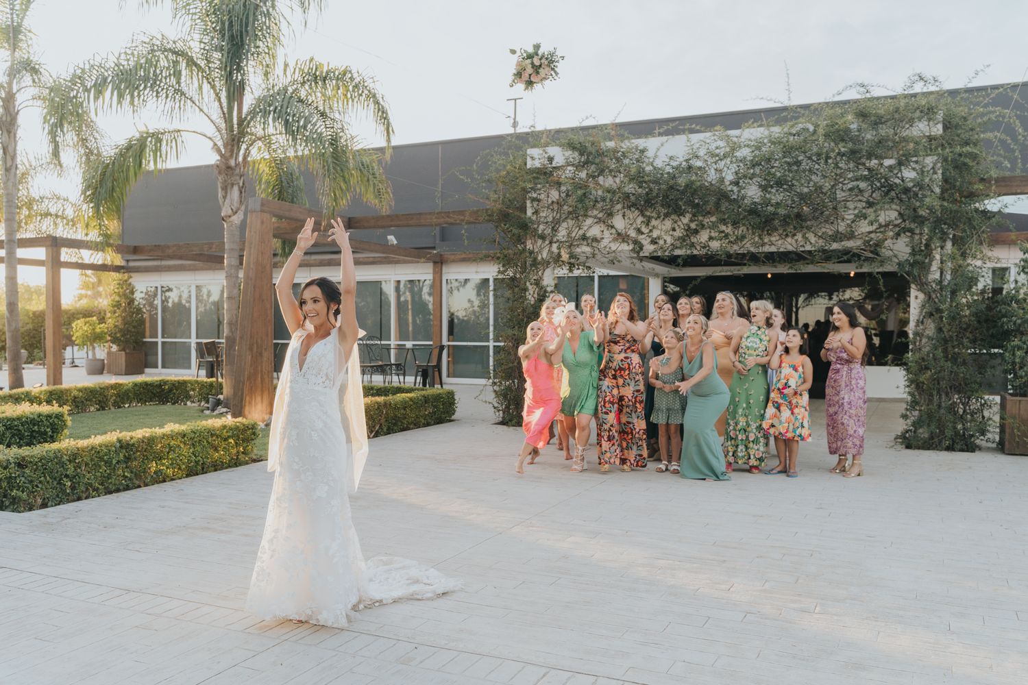 A bride in a flowing white gown poses while a group in colorful dresses cheers from behind at an outdoor venue.