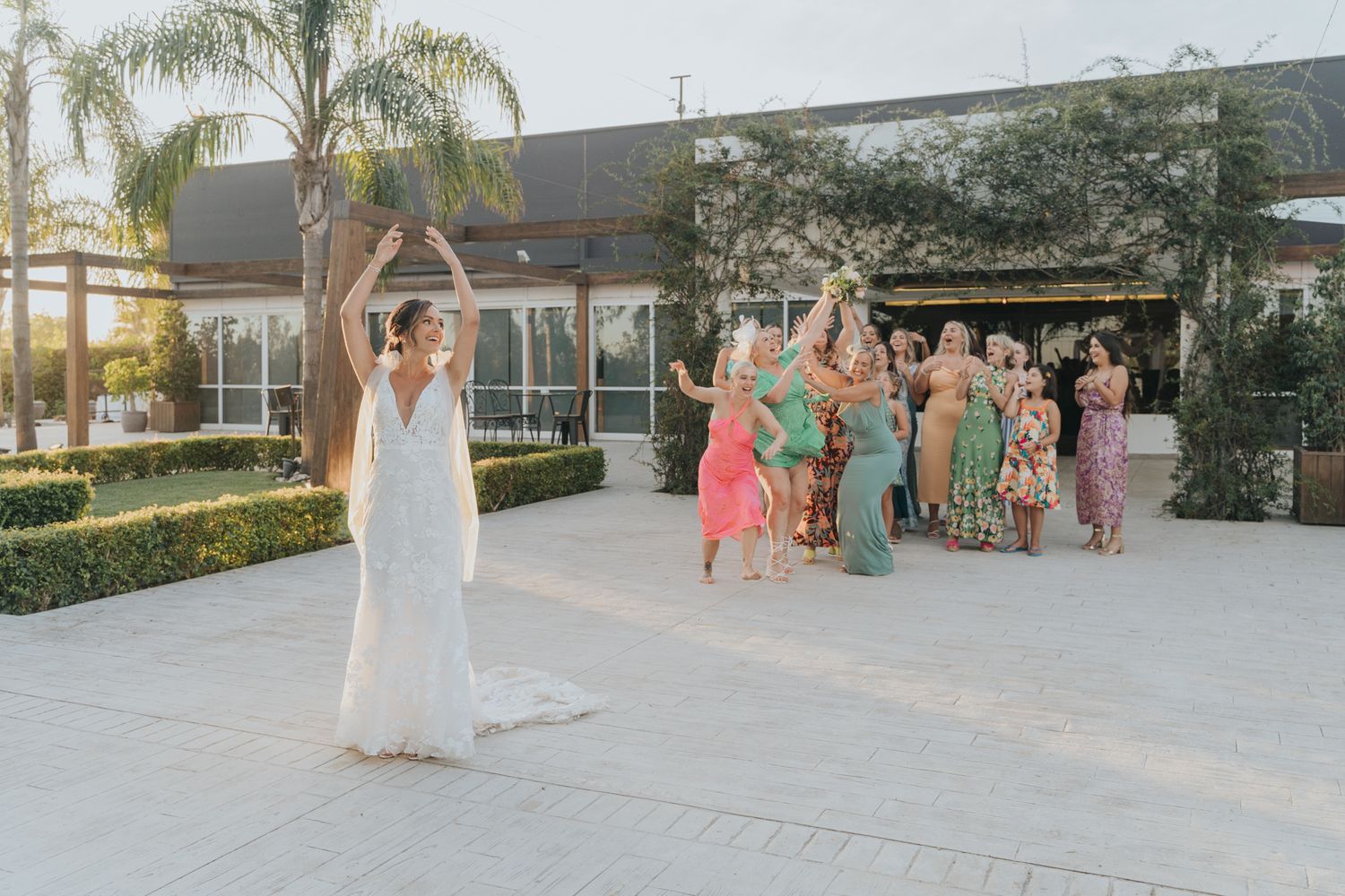 Wedding bouquet toss scene at an outdoor venue with bridesmaids in coral and sage dresses.