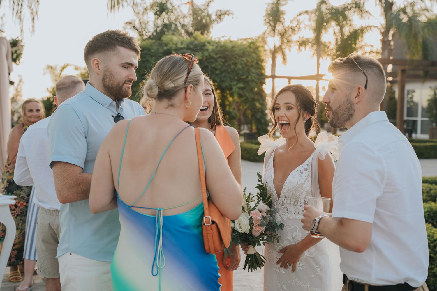 Wedding guests mingle during sunset at an outdoor reception with palm trees in the background.
