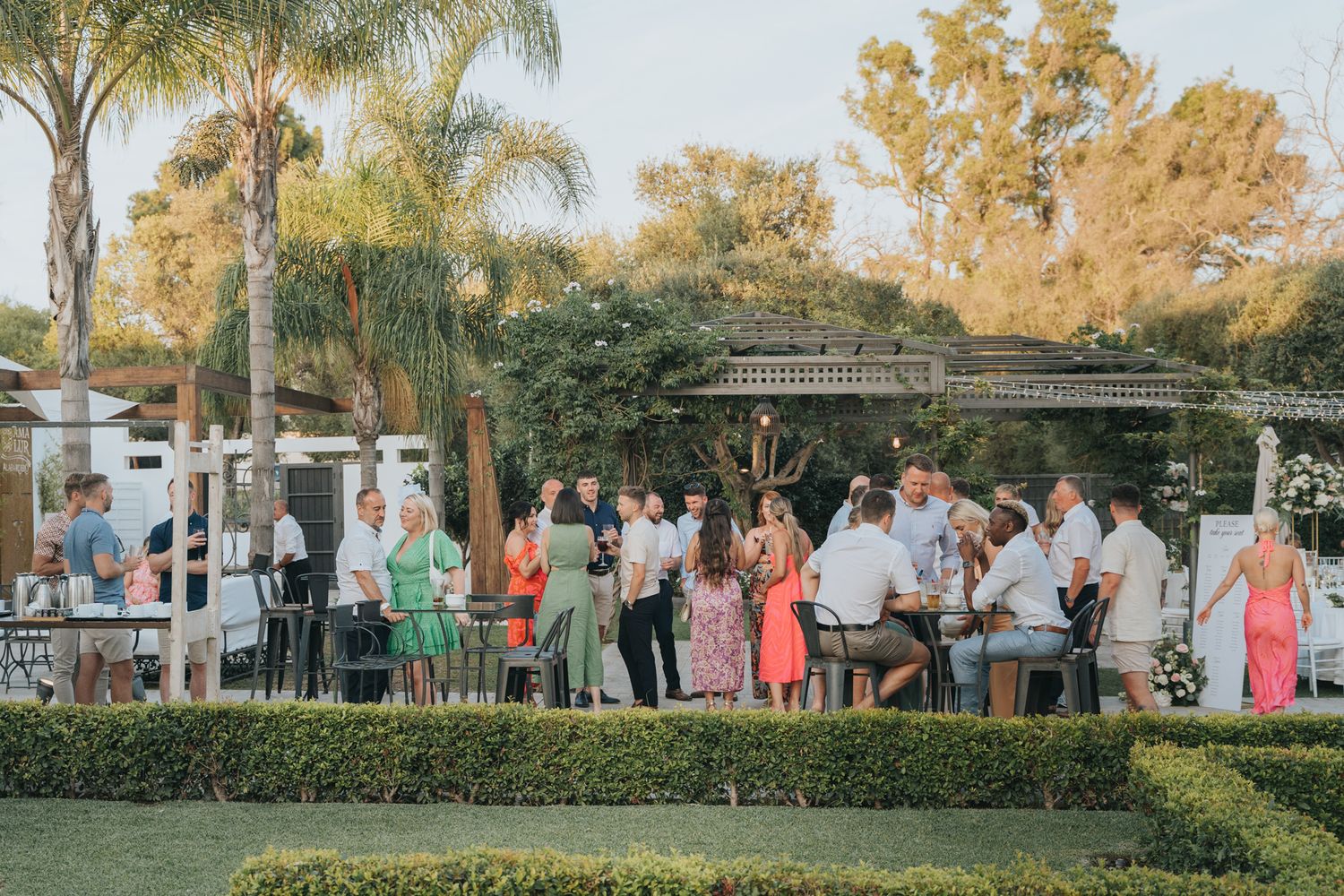 Wedding guests mingle at an elegant outdoor reception with palm trees and decorative lighting.