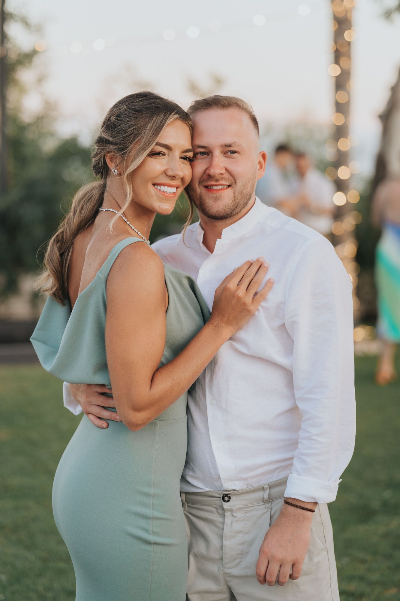 A couple embraces outdoors wearing formal attire at a garden party with twinkling lights in the background.