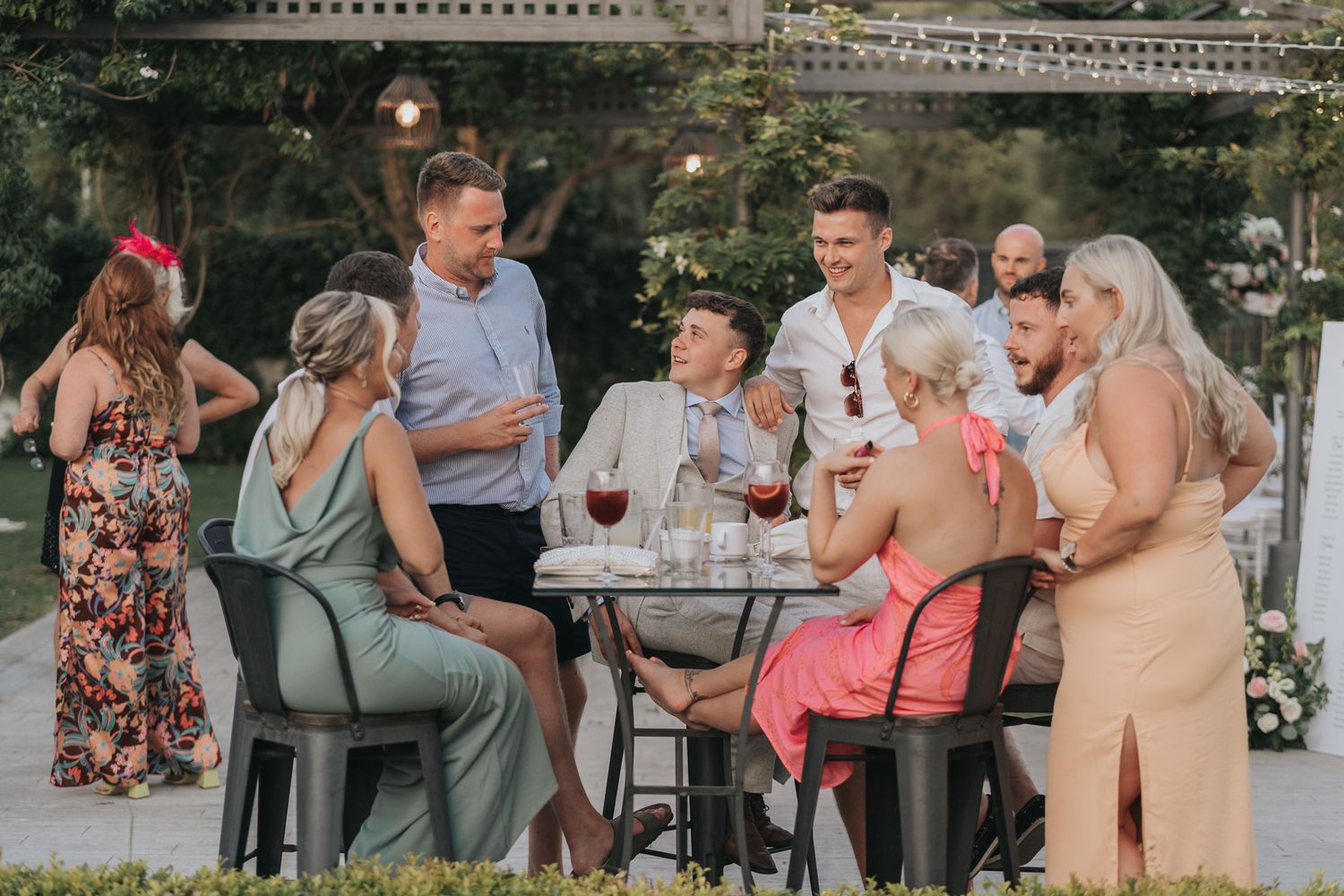 A group gathers around an outdoor table for drinks and conversation during a summer celebration.