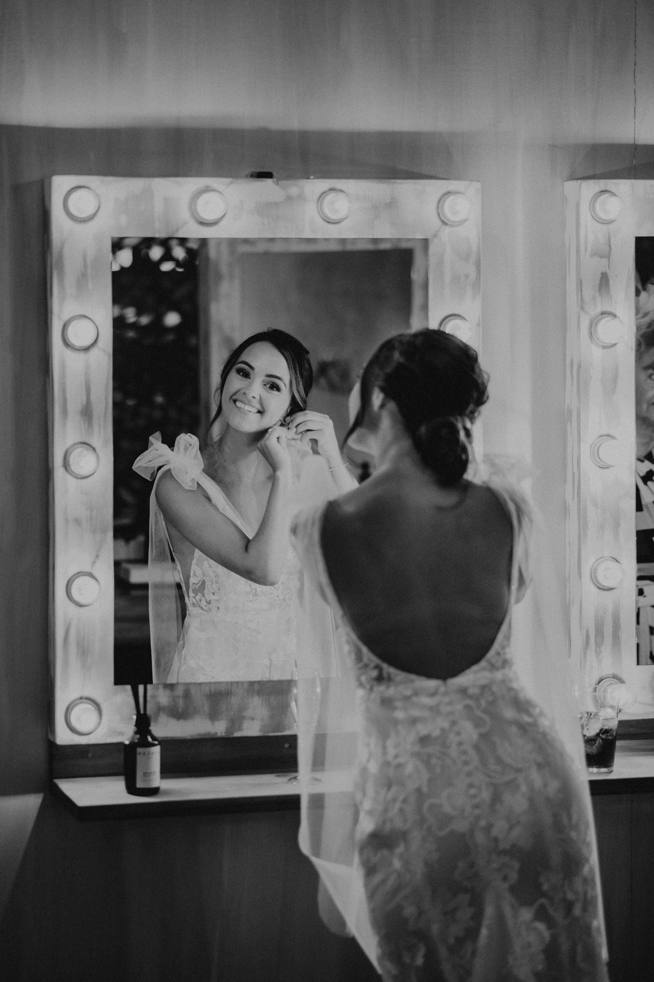 A woman in a lace dress looks at her reflection while getting ready in a dressing room with vanity lights.