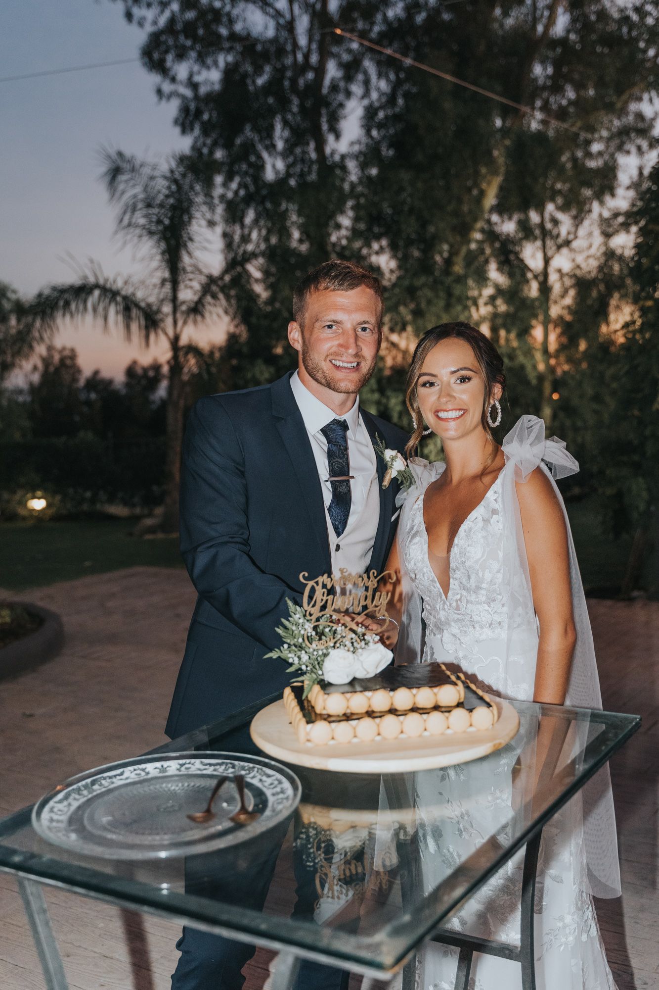 A couple cuts their wedding cake together at dusk with decorative flowers atop a glass-topped dessert table.