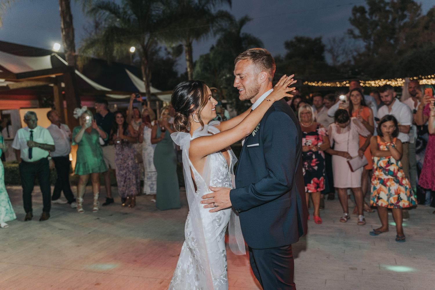 A couple shares their first dance at an evening wedding reception while guests watch from the dance floor.