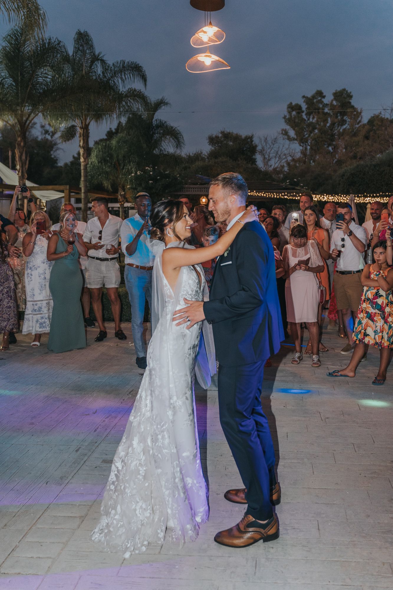 Evening wedding dance under string lights with guests gathered around the outdoor dance floor.