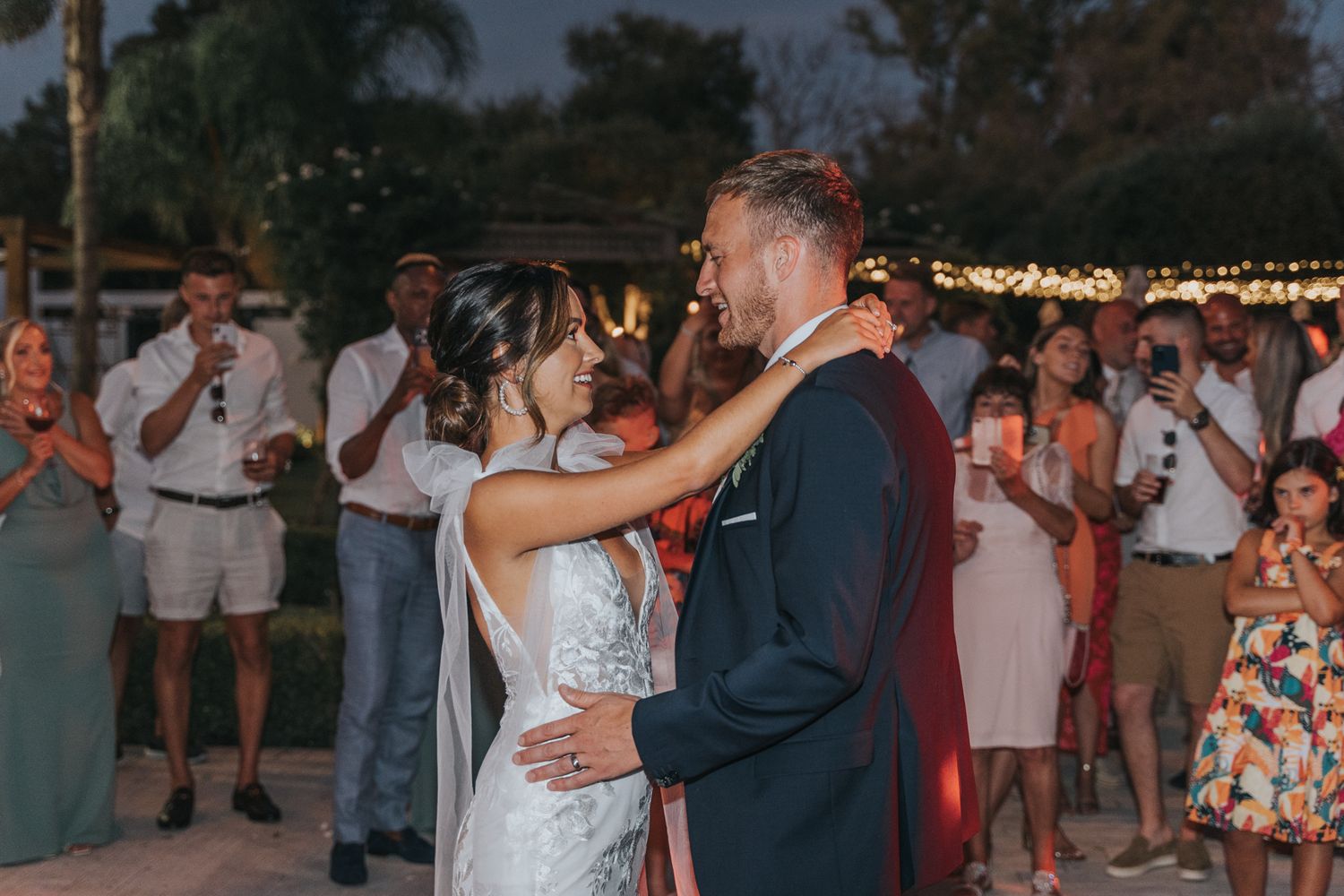 A couple shares their first dance at an outdoor wedding reception as guests look on under string lights at dusk.