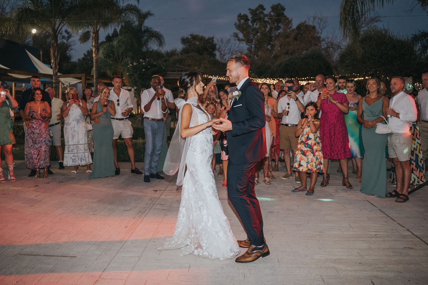 A couple shares their first dance at an outdoor evening wedding reception while guests look on from the sidelines.