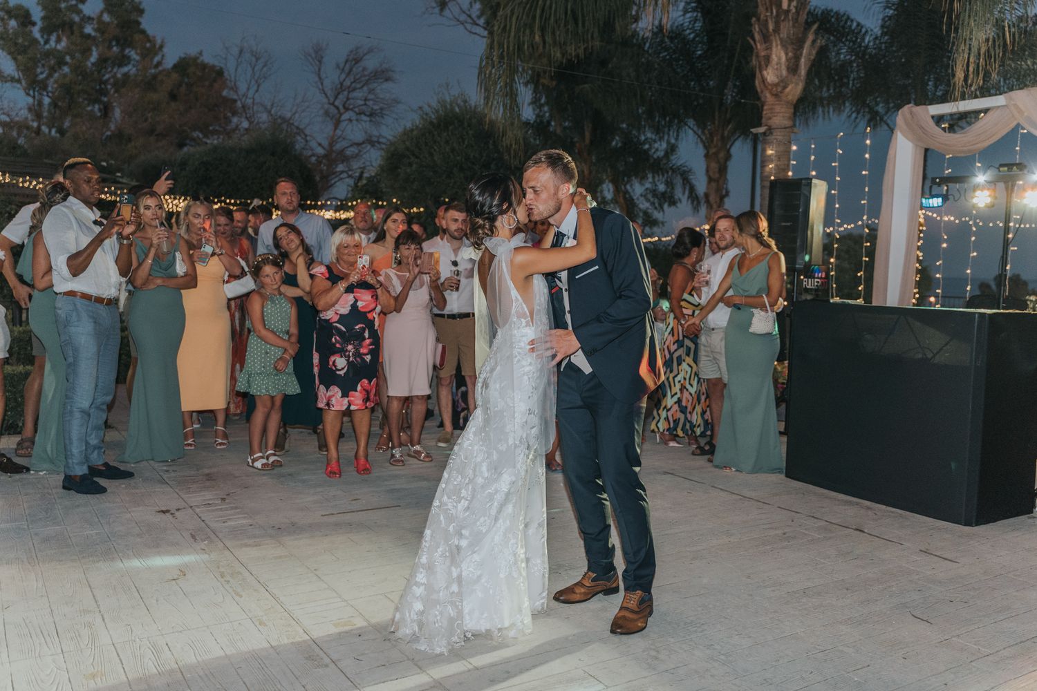 Couple shares their first dance at an outdoor evening wedding reception with guests watching and palm trees in background.