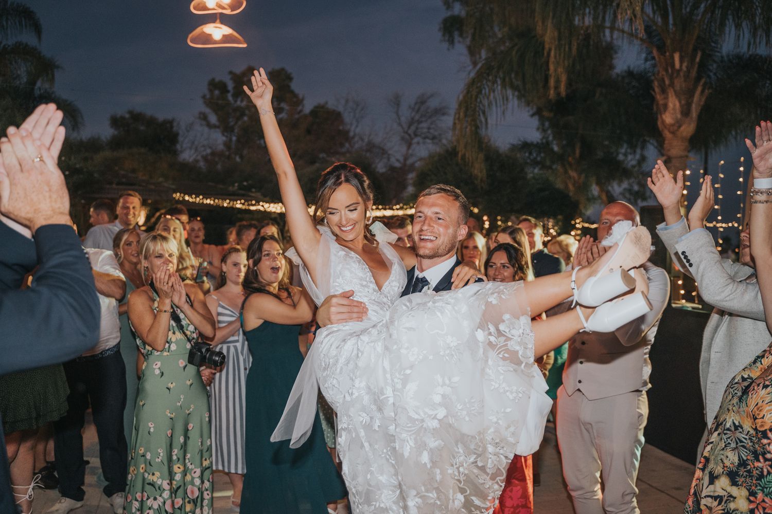 Joyous celebration at an outdoor evening wedding reception with dancing and cheering guests under string lights.