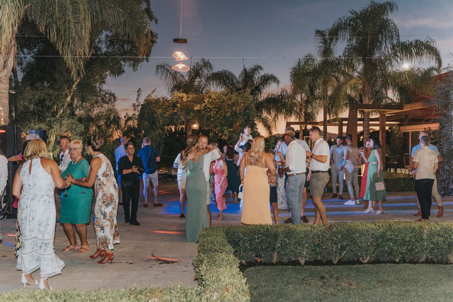 Wedding reception guests mingling on an outdoor patio decorated with palm trees and ambient lighting.