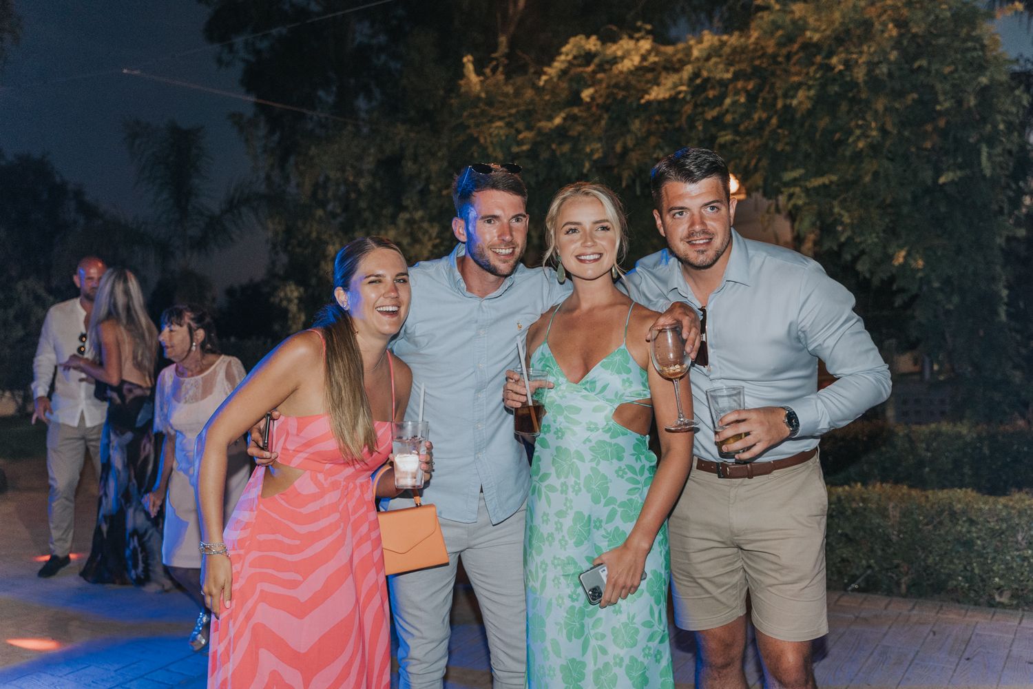 Wedding guests in summer attire pose together at an outdoor evening celebration.