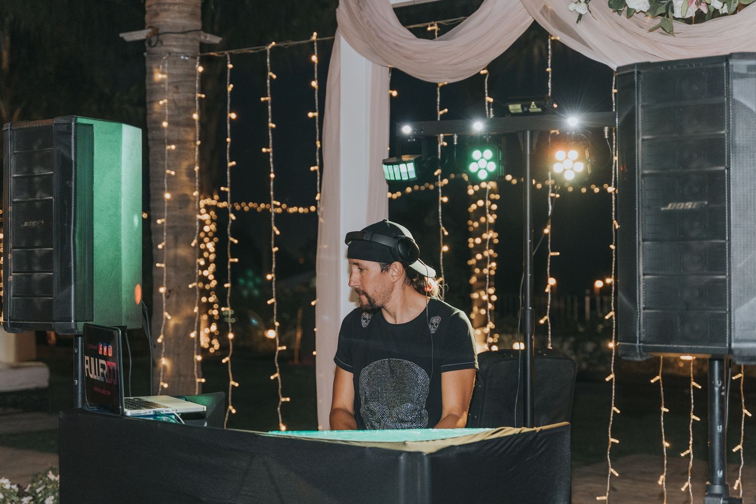 DJ performs behind illuminated booth with hanging fairy lights and white drapes creating an enchanting atmosphere at event.