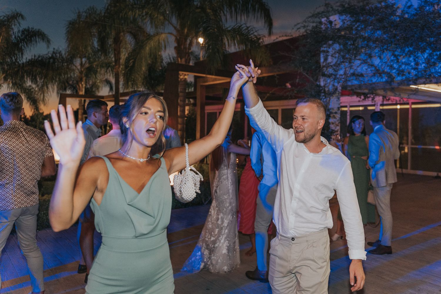Wedding guests dance enthusiastically at an outdoor reception with palm trees in the background at dusk.