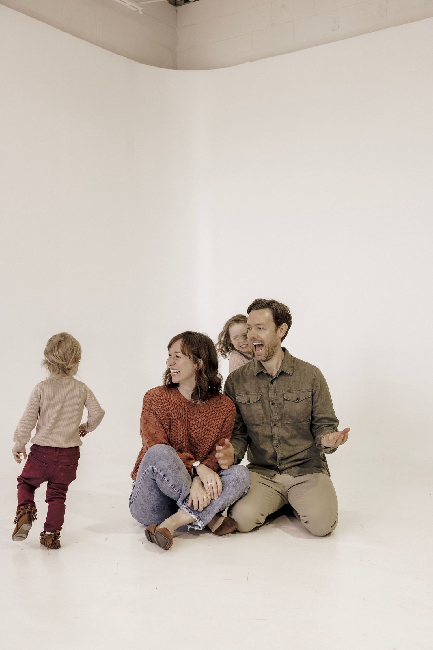 Family of 4 with two young children laughing as son runs around the room