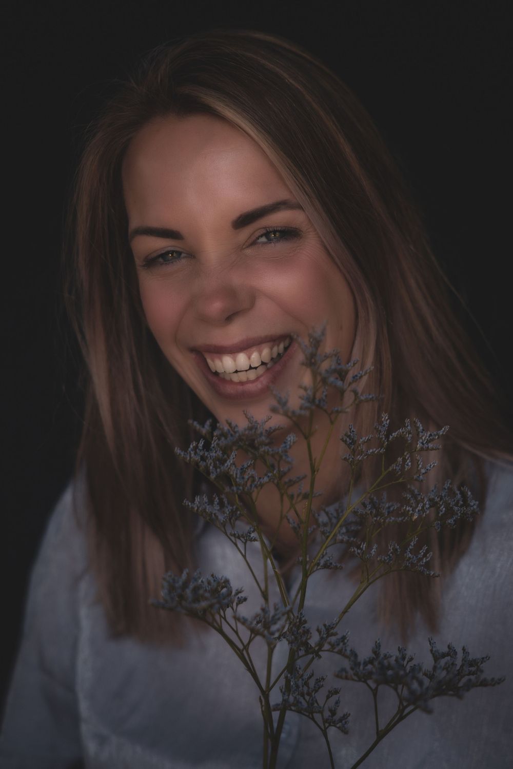 Portrait with delicate purple flowers in foreground showing joyful expression against dark background.