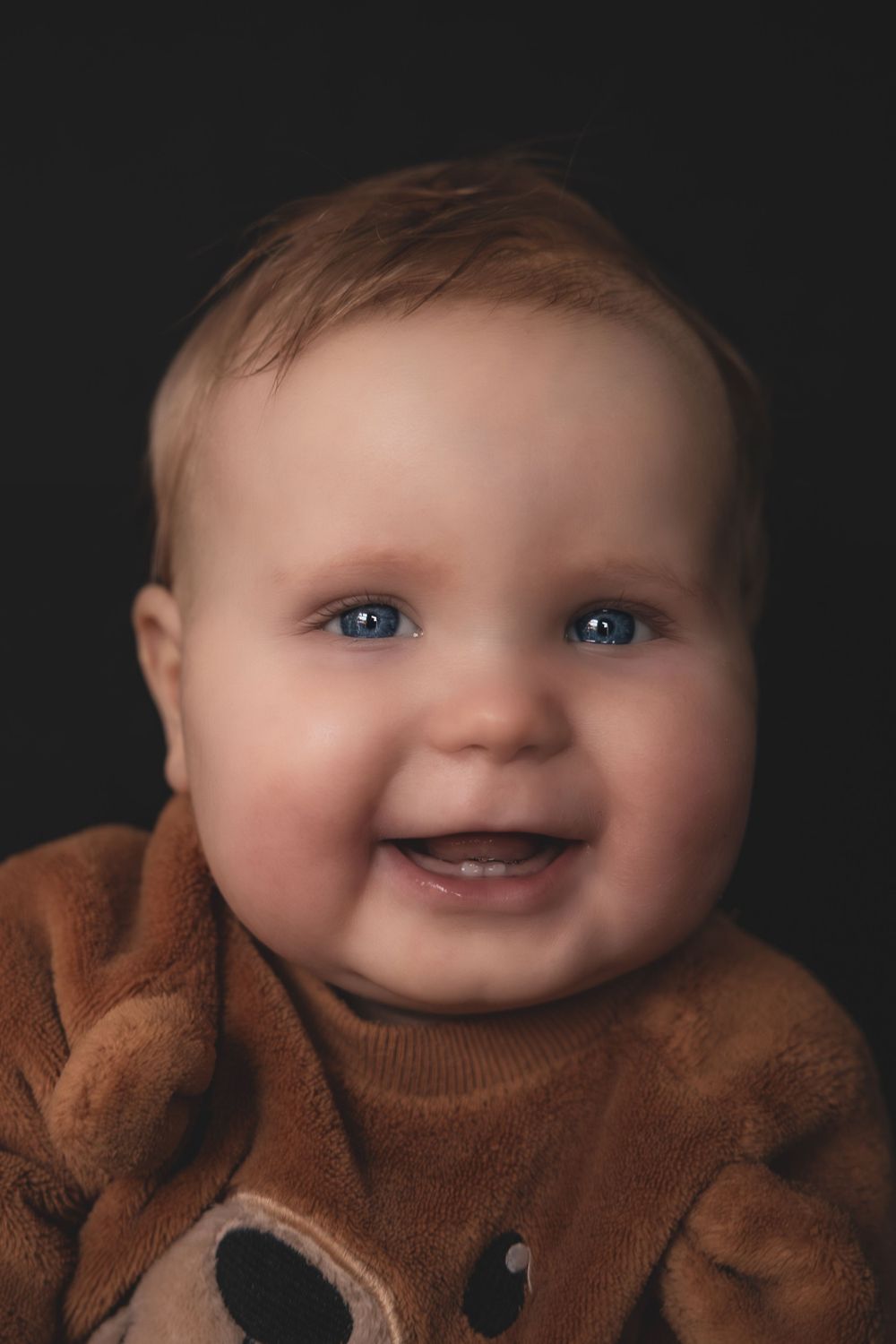 Adorable baby wearing brown outfit smiles brightly against dark background, showing bright blue eyes.