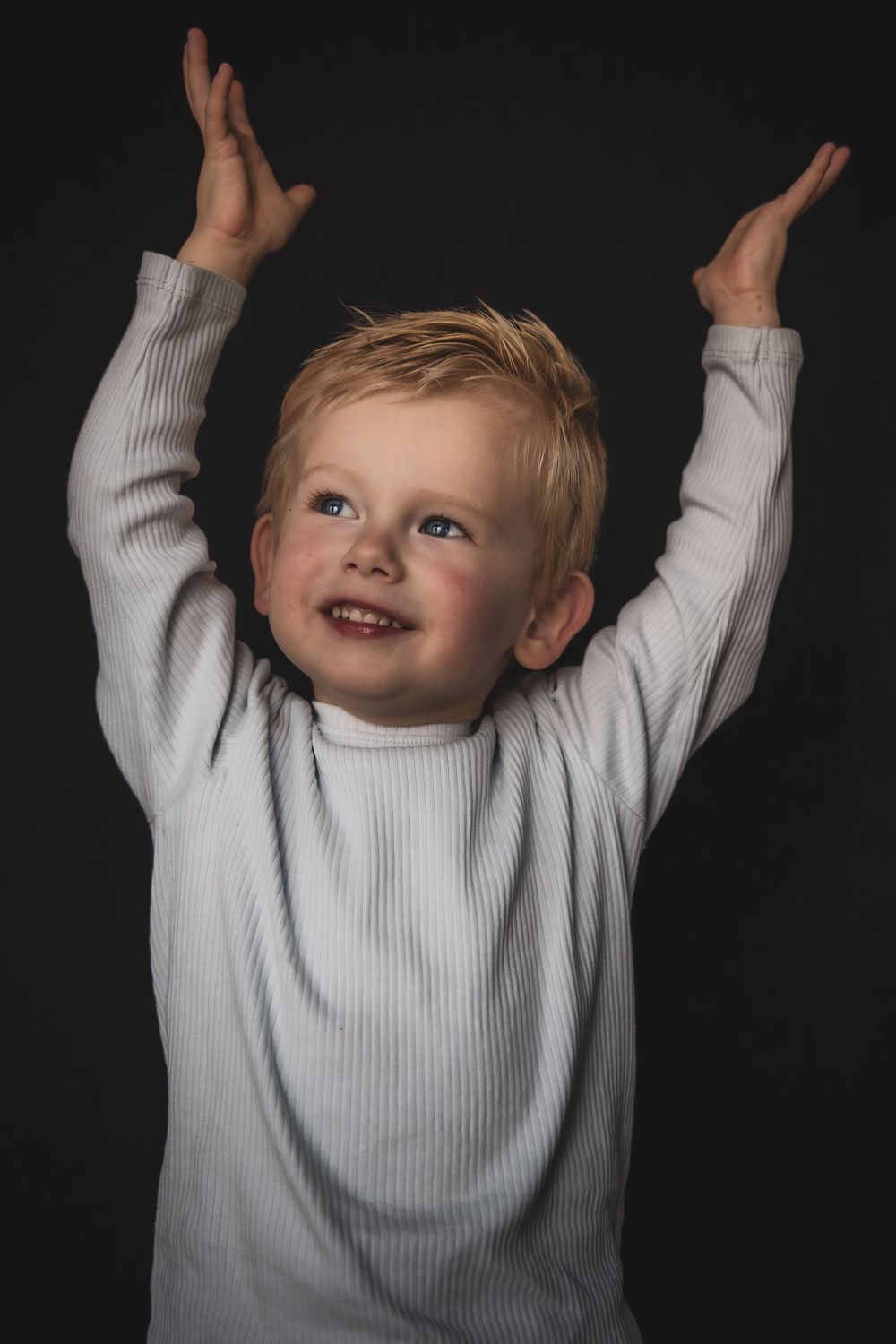 A joyful child in a white sweater raises their arms up against a dark background in a sequence of photos.
