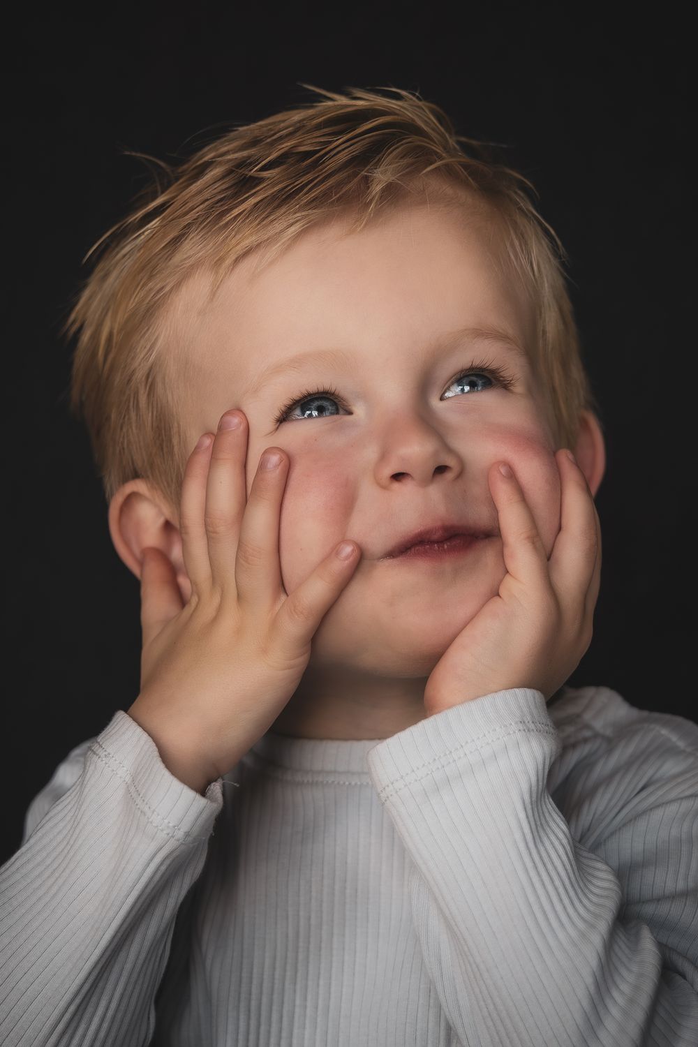 A child in a white sweater makes playful expressions while covering their face against a dark background.