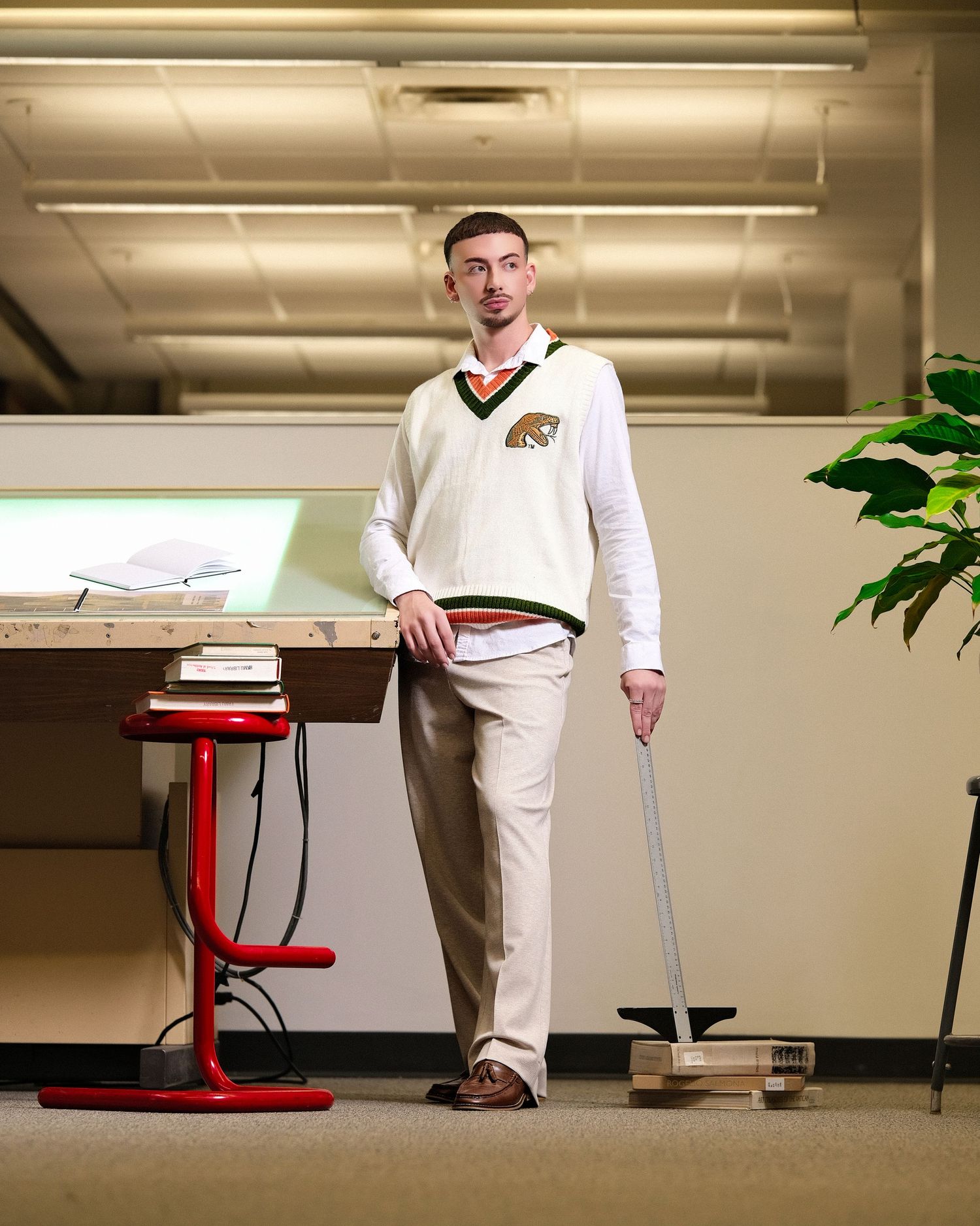 Person in white sweater, khaki pants, and glasses leans against a podium in an indoor office setting with fluorescent lights.