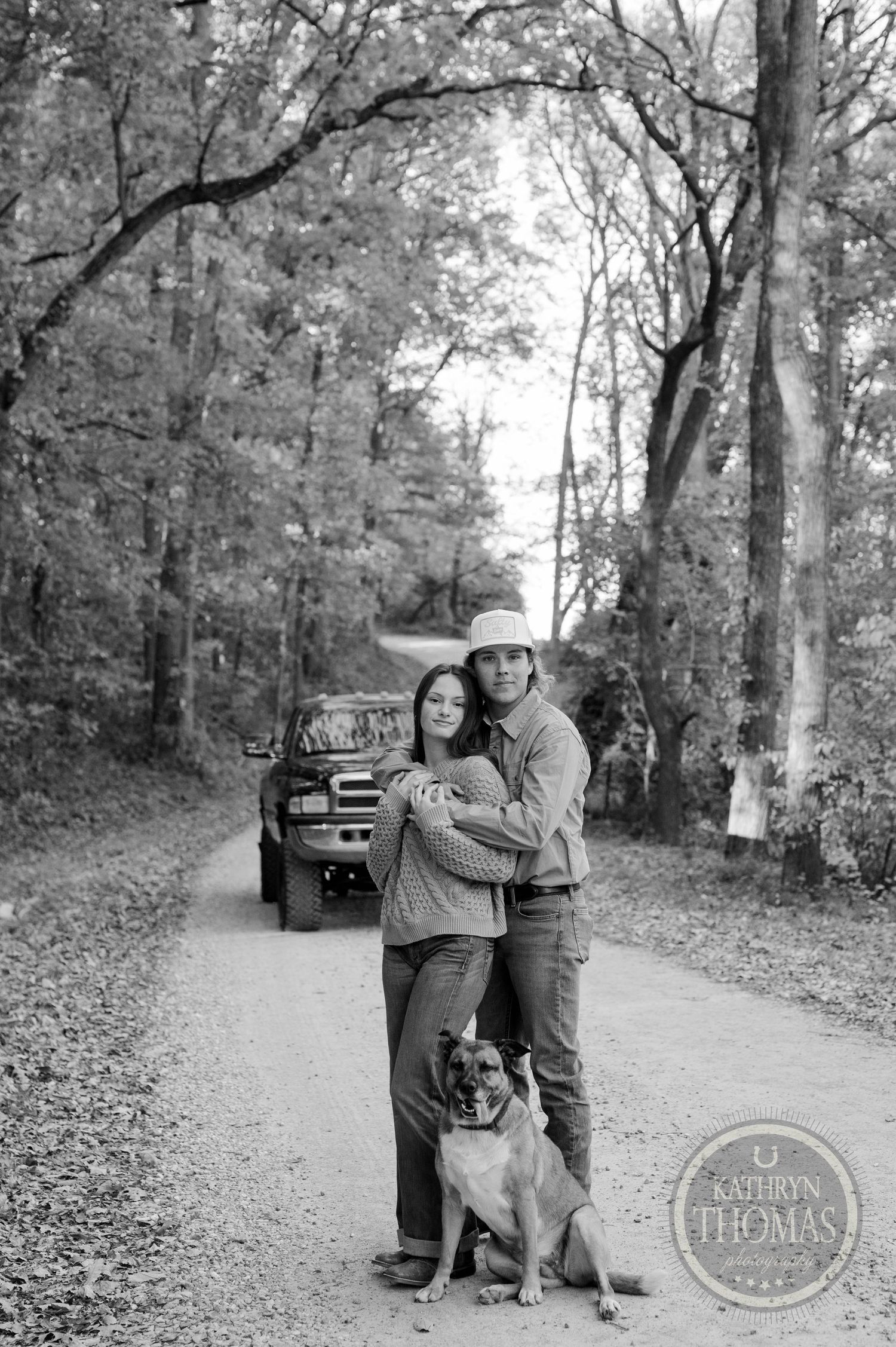 A couple embraces with their dog on a wooded dirt road with a pickup truck in the background.