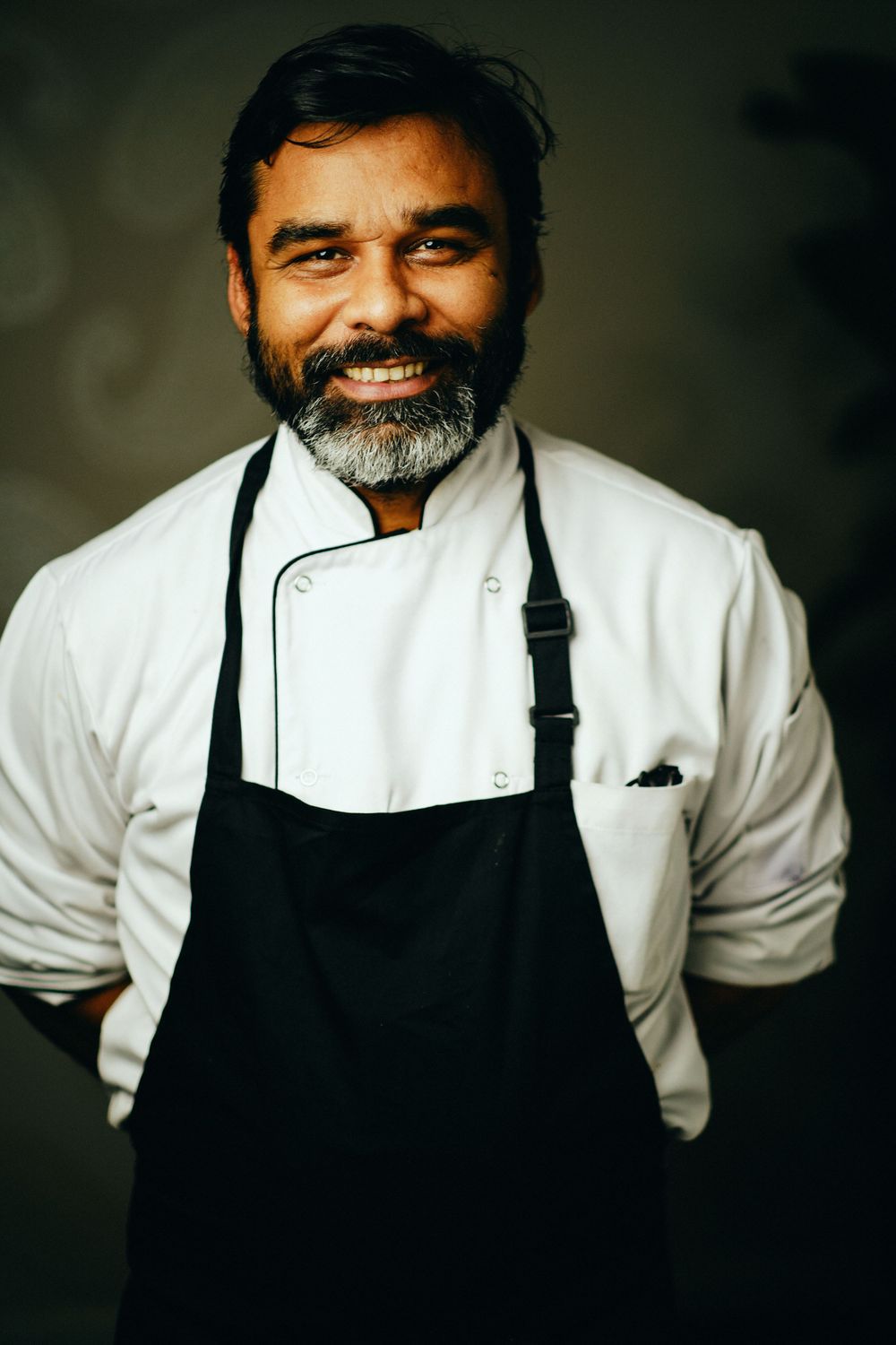 Chef in white coat and black apron smiles warmly against dark background.