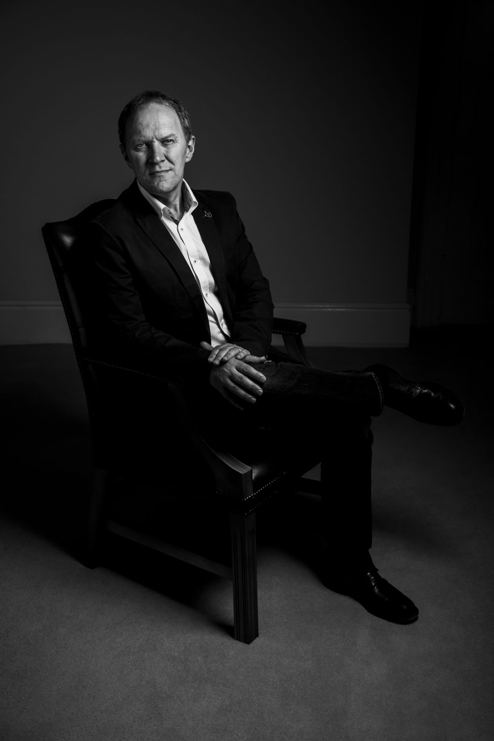 Black and white portrait in dramatic lighting showing seated figure in formal attire.