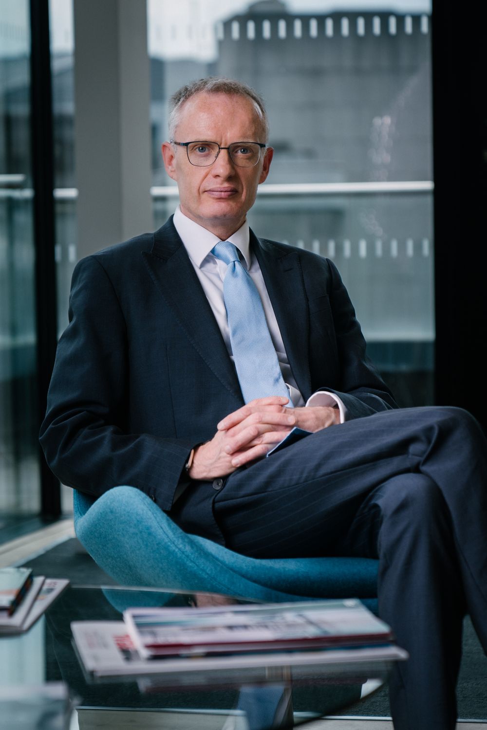 Professional businessman in a navy suit and glasses sits in a modern office setting.