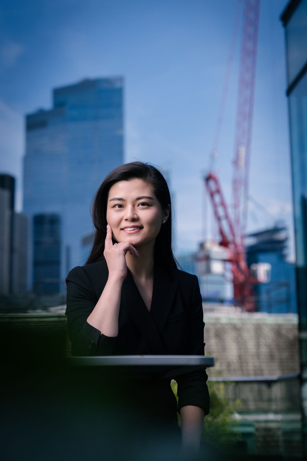 Professional in black attire poses thoughtfully against urban backdrop with construction crane.
