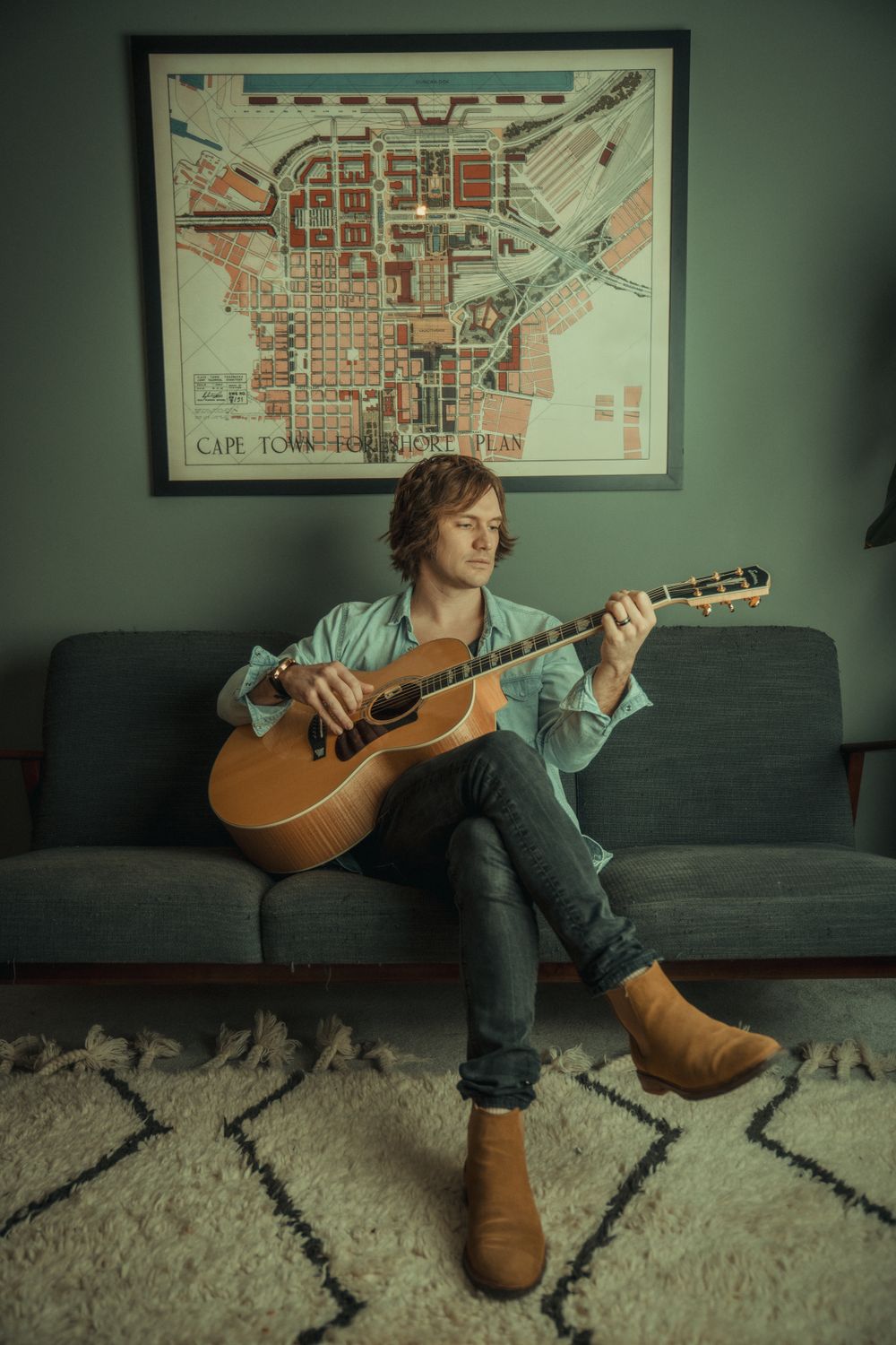Musician plays acoustic guitar while sitting on couch beneath vintage city map.