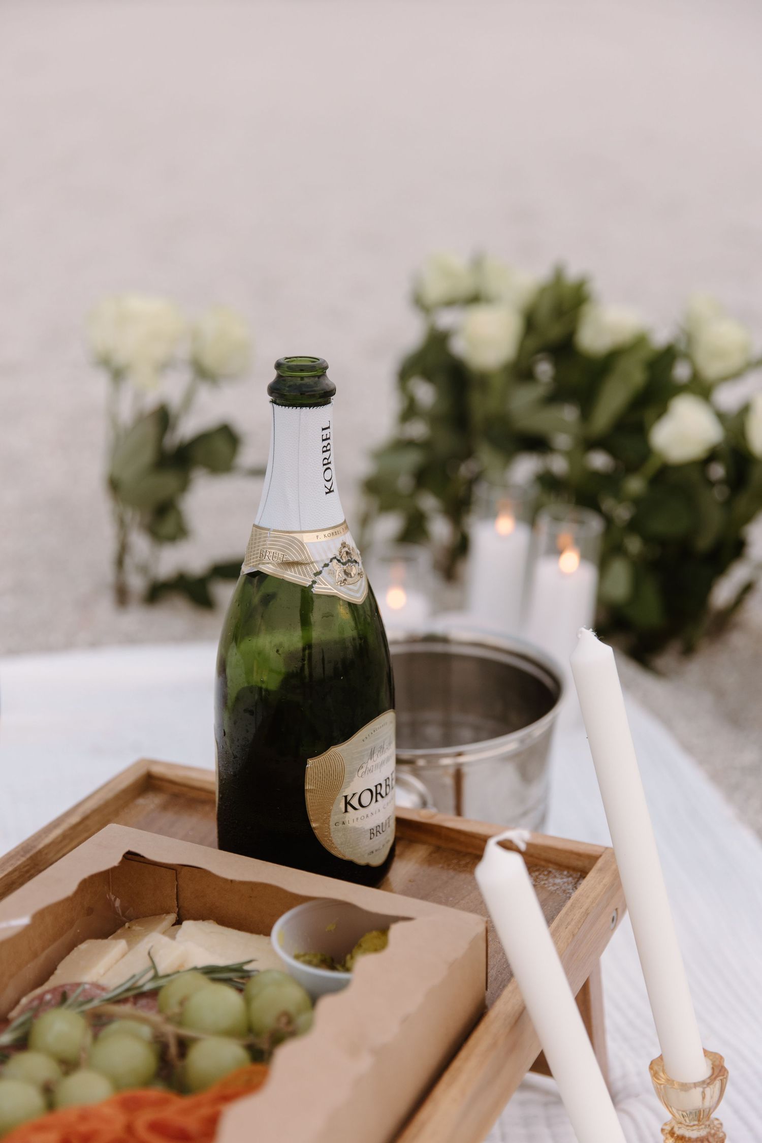 Elegant charcuterie board with champagne, flowers, and assorted snacks displayed on white tablecloth.