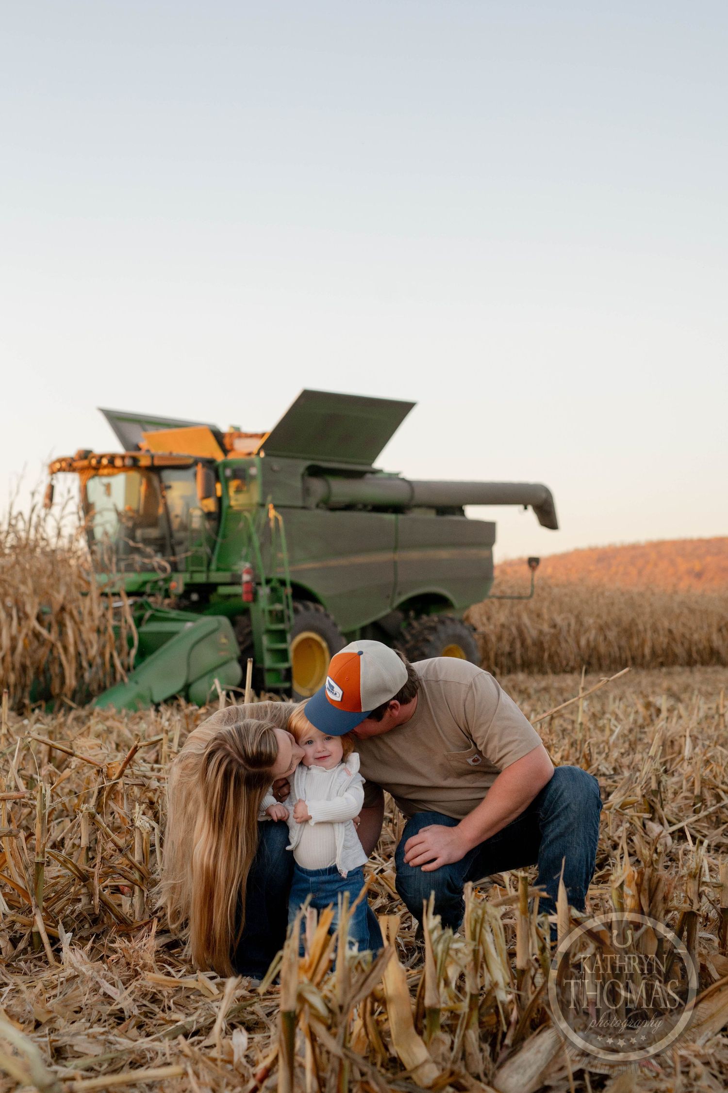Family shares intimate moment in cornfield with John Deere combine during golden hour.