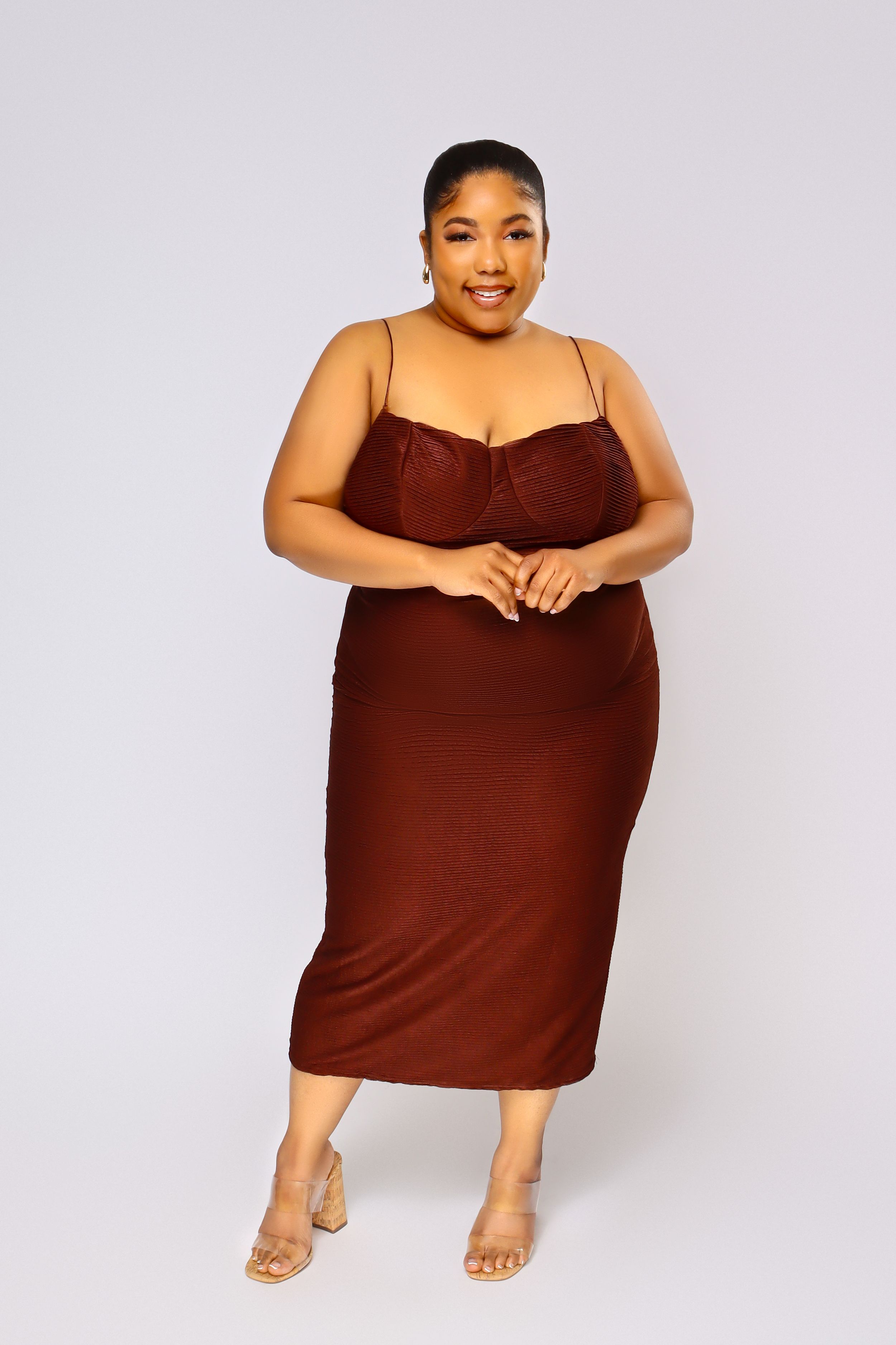 Curvy model elegantly poses in a brown fitted midi dress and nude sandals against light grey background.