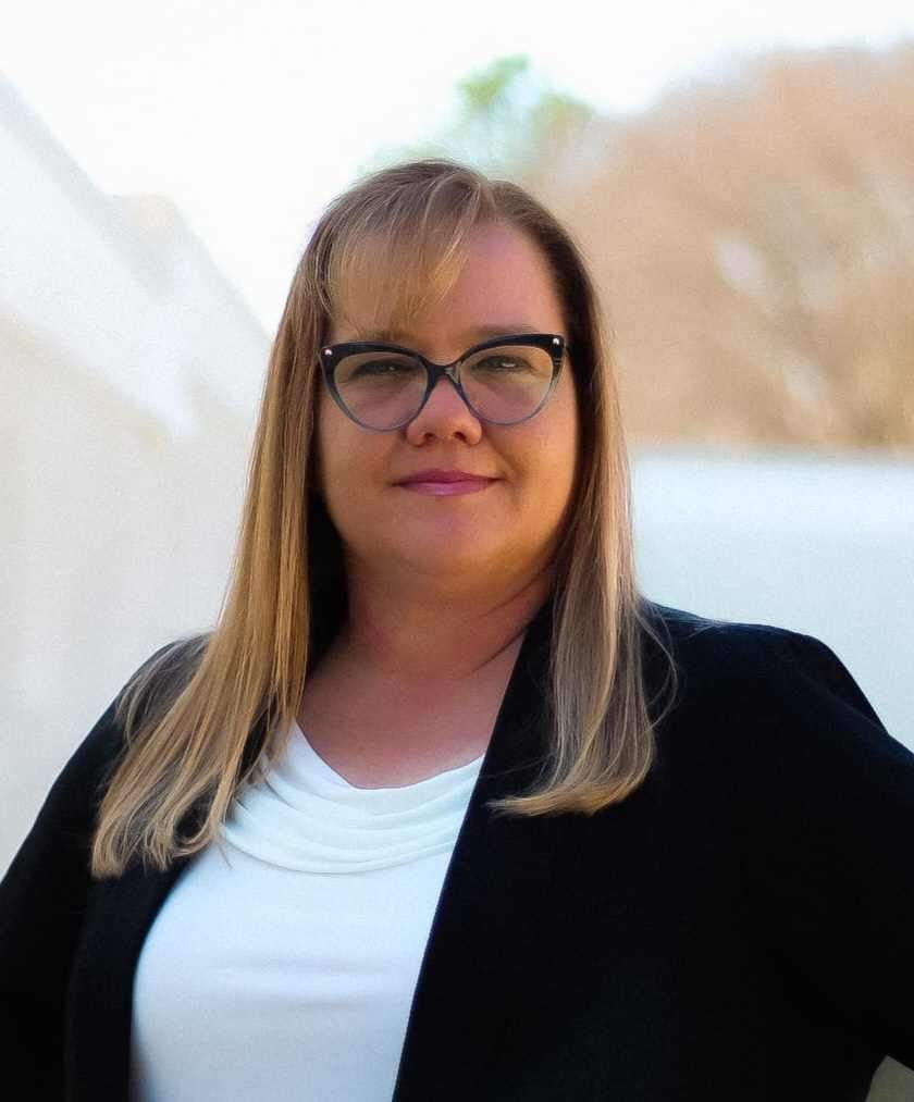 Professional headshot of person wearing glasses and black blazer against blurred background.