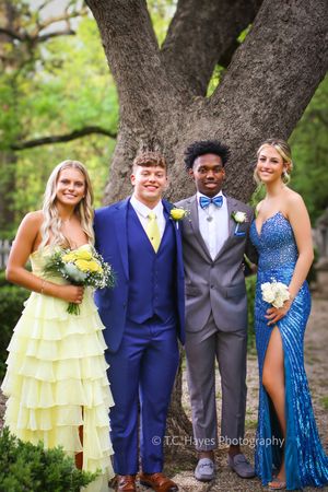 Four students pose in formal attire next to a large tree trunk at prom, wearing yellow, blue and sparkling dresses and suits.