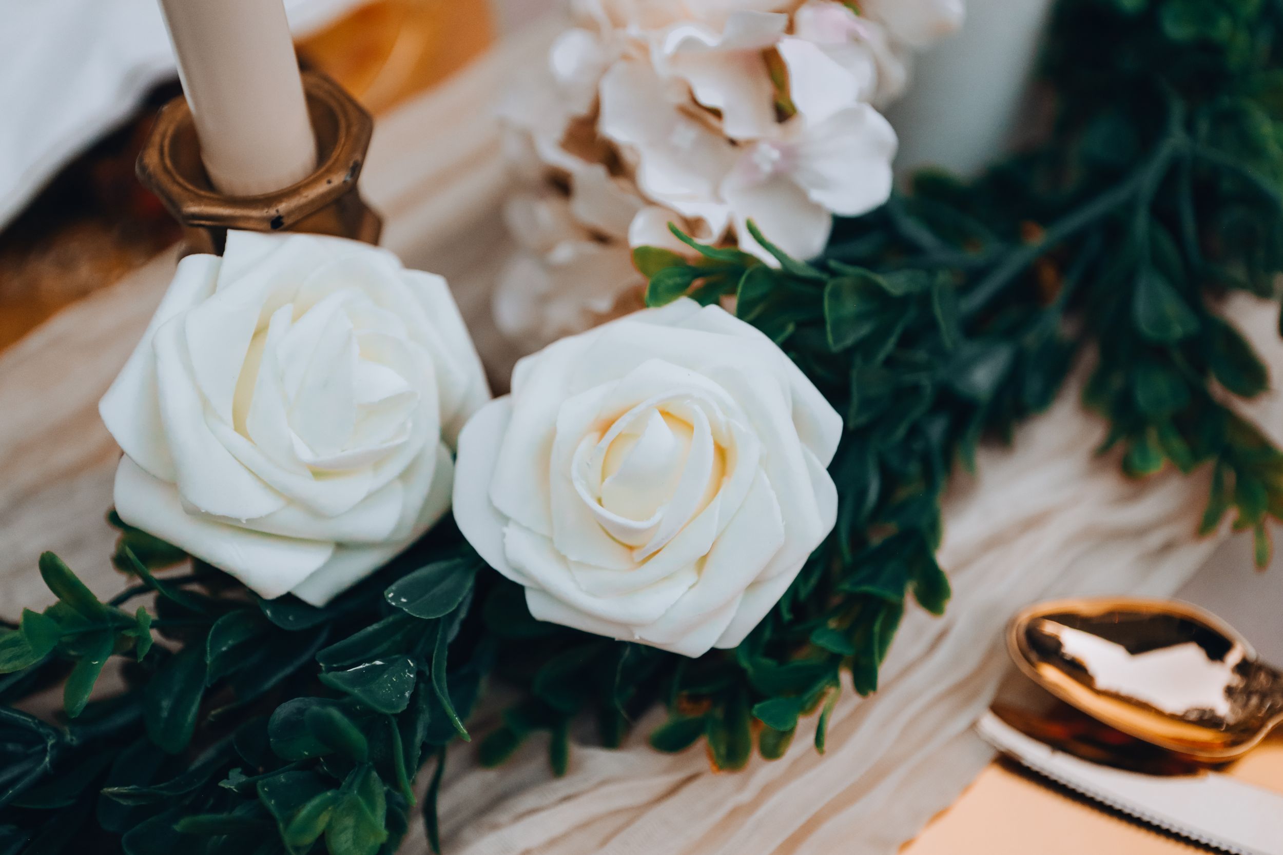 Two white roses with green foliage arranged next to a candle on a light surface.