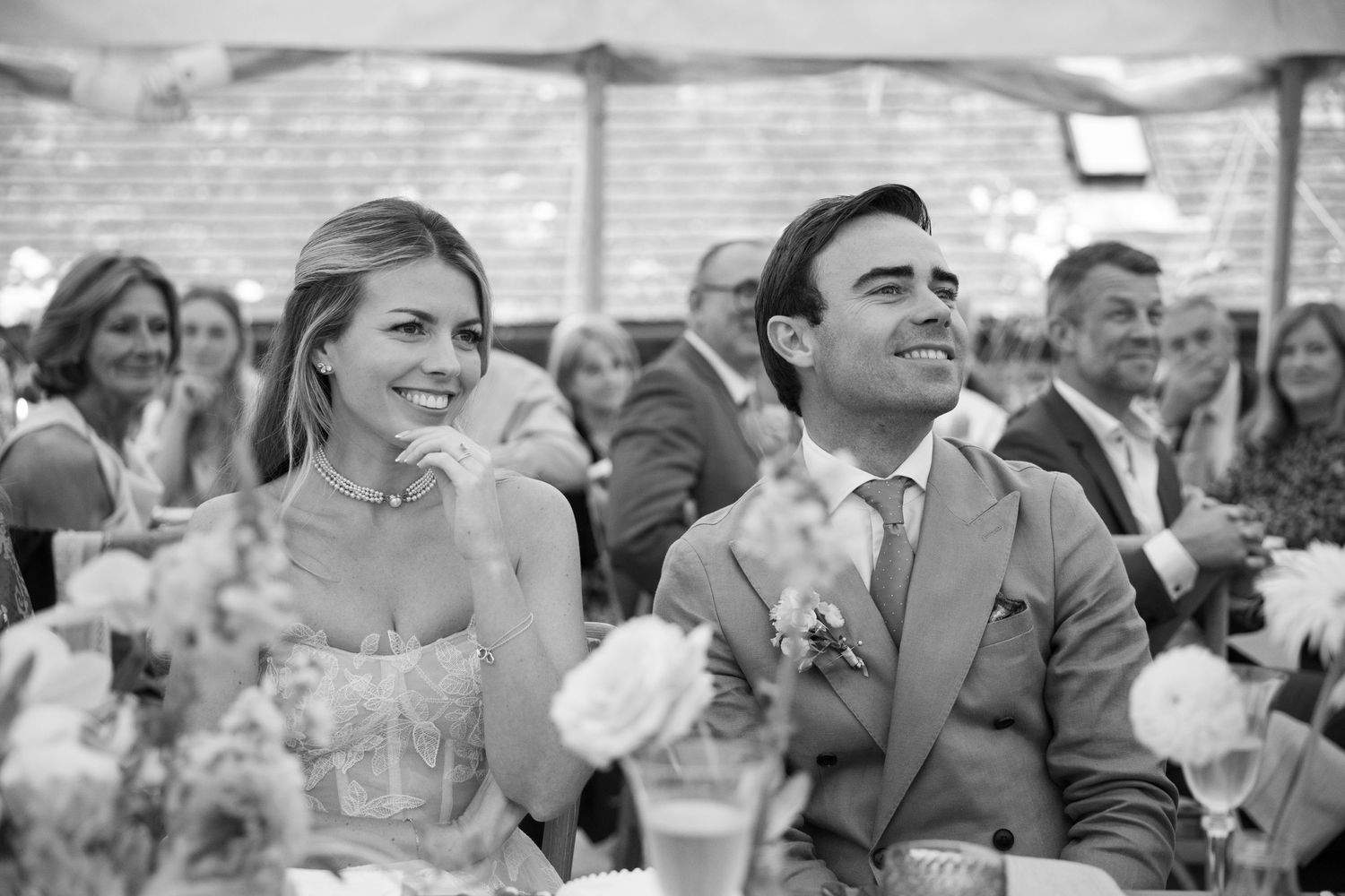 A black and white photo of a seated couple at a wedding reception laughing among guests.