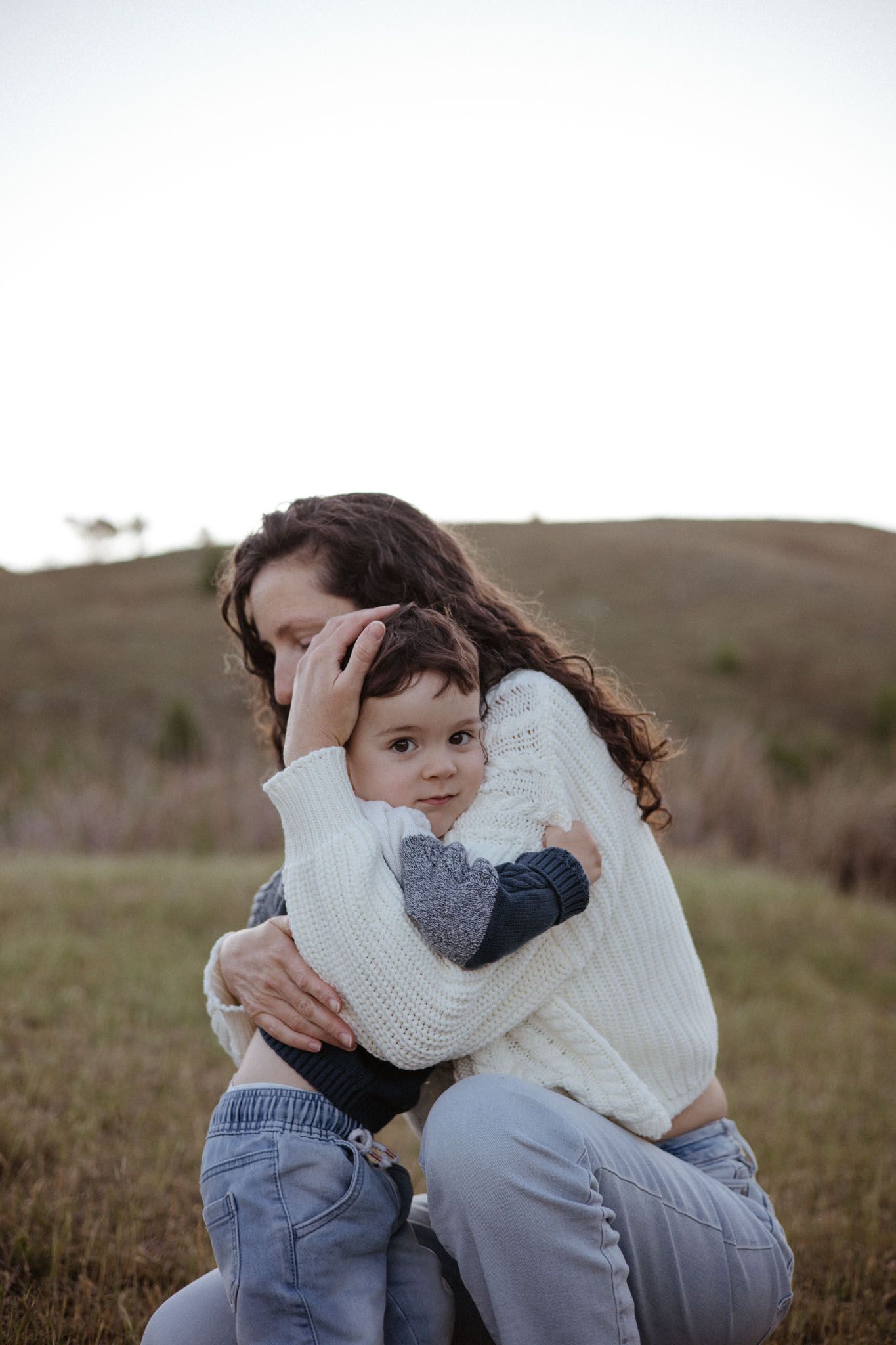 A tender embrace between parent and child wearing white sweaters outdoors in a natural field setting at dusk.