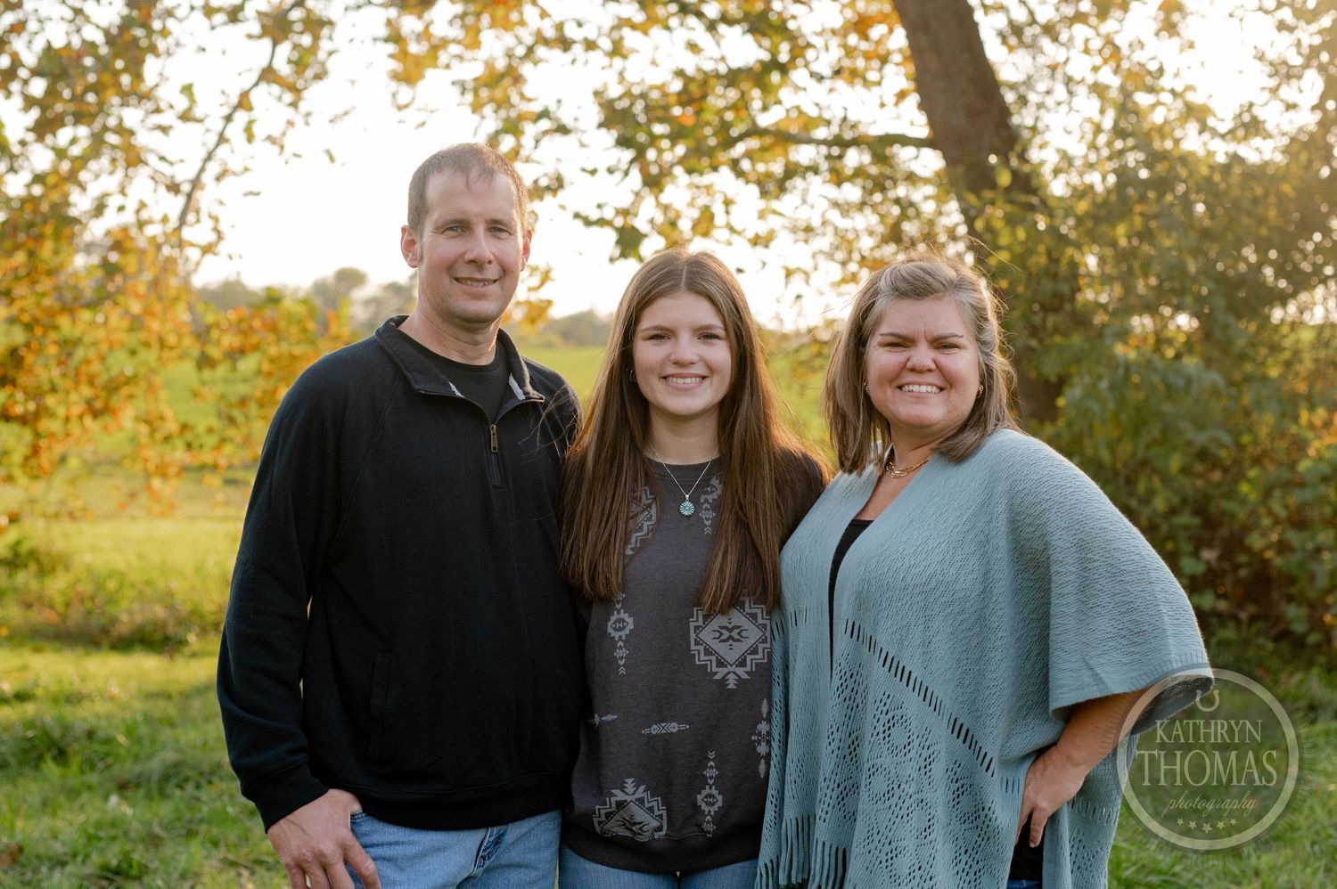 Three people smile together outdoors during golden hour with autumn trees in background.