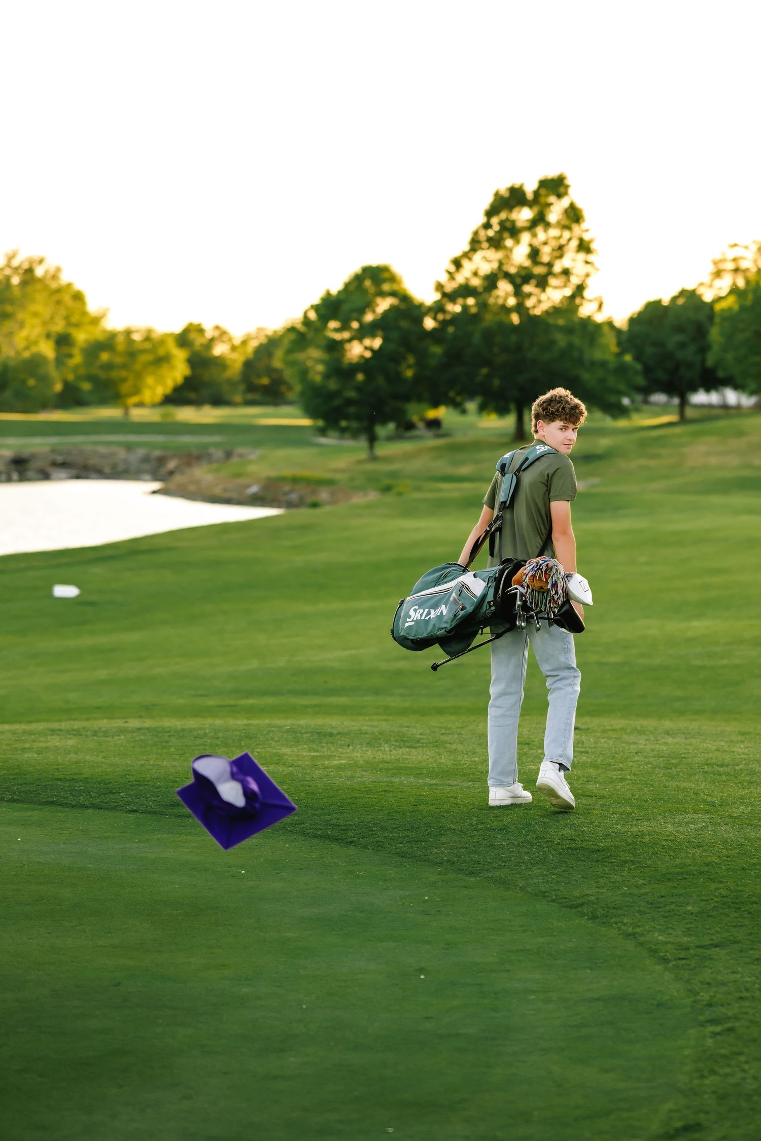 A golfer walks across a lush green course at sunset carrying a golf bag and gear.