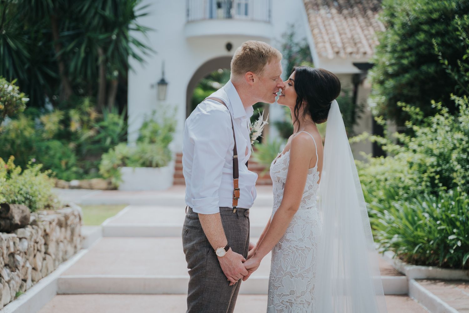 A romantic moment between newlyweds holding hands on the garden steps of a white stucco Mediterranean villa.
