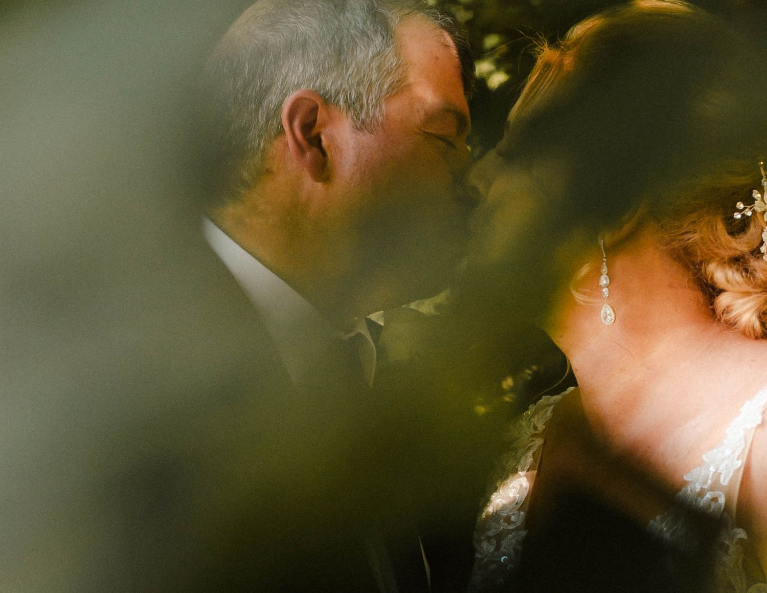 A romantic wedding moment captured through leaves and soft lighting at an outdoor ceremony.