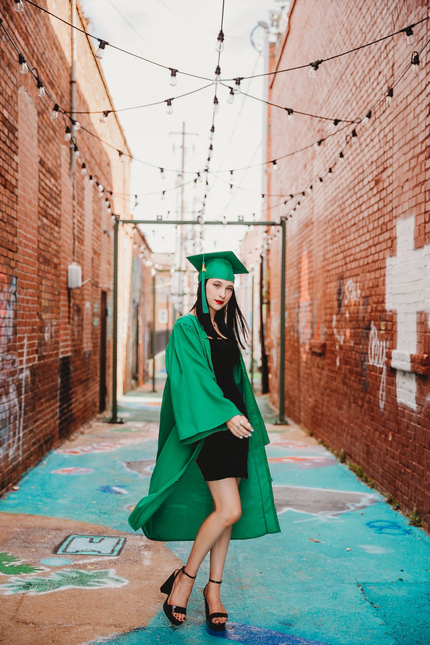 Graduate posing in emerald green cap and gown in an urban alley with brick walls and string lights overhead.