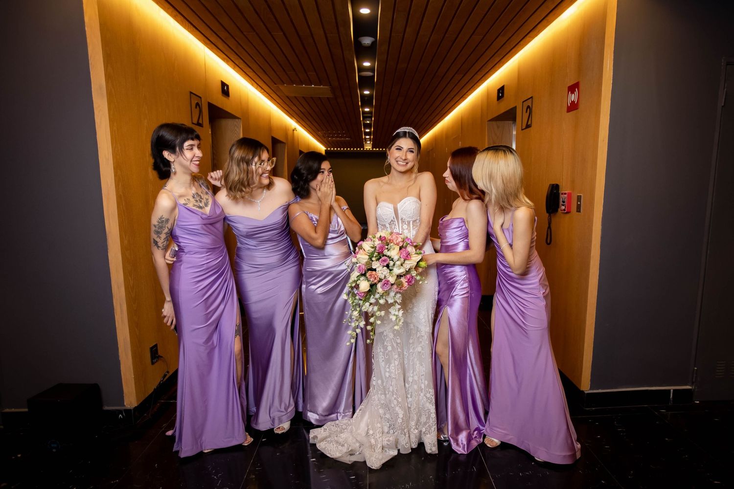 Bridal party in matching purple gowns posing in an elegantly lit hotel hallway.