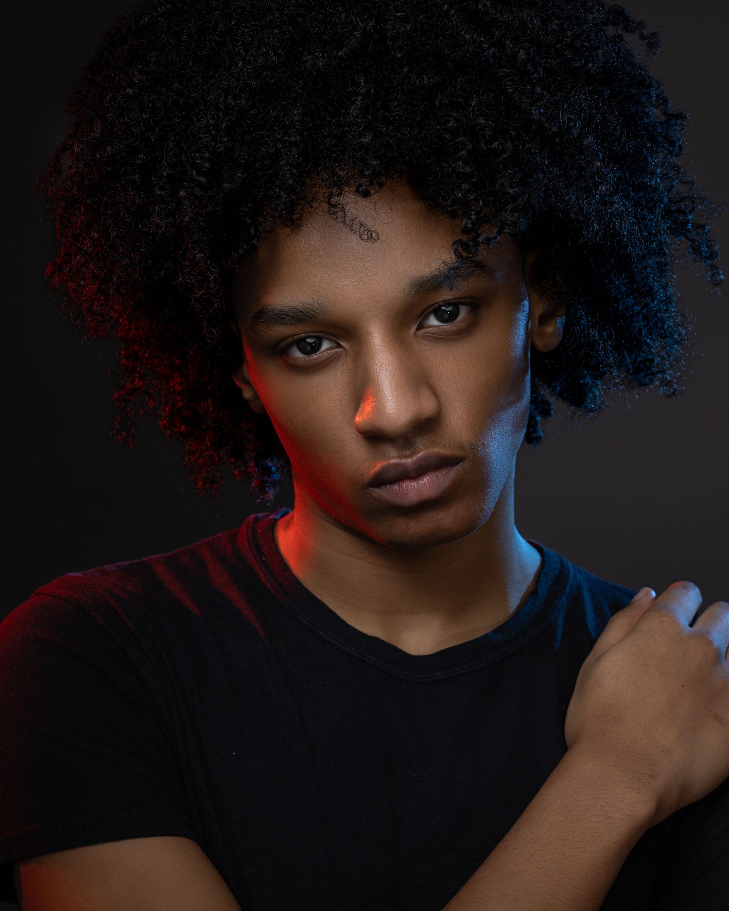 Portrait photo with dramatic red and blue lighting showing person in black shirt with natural curly hair against dark background.