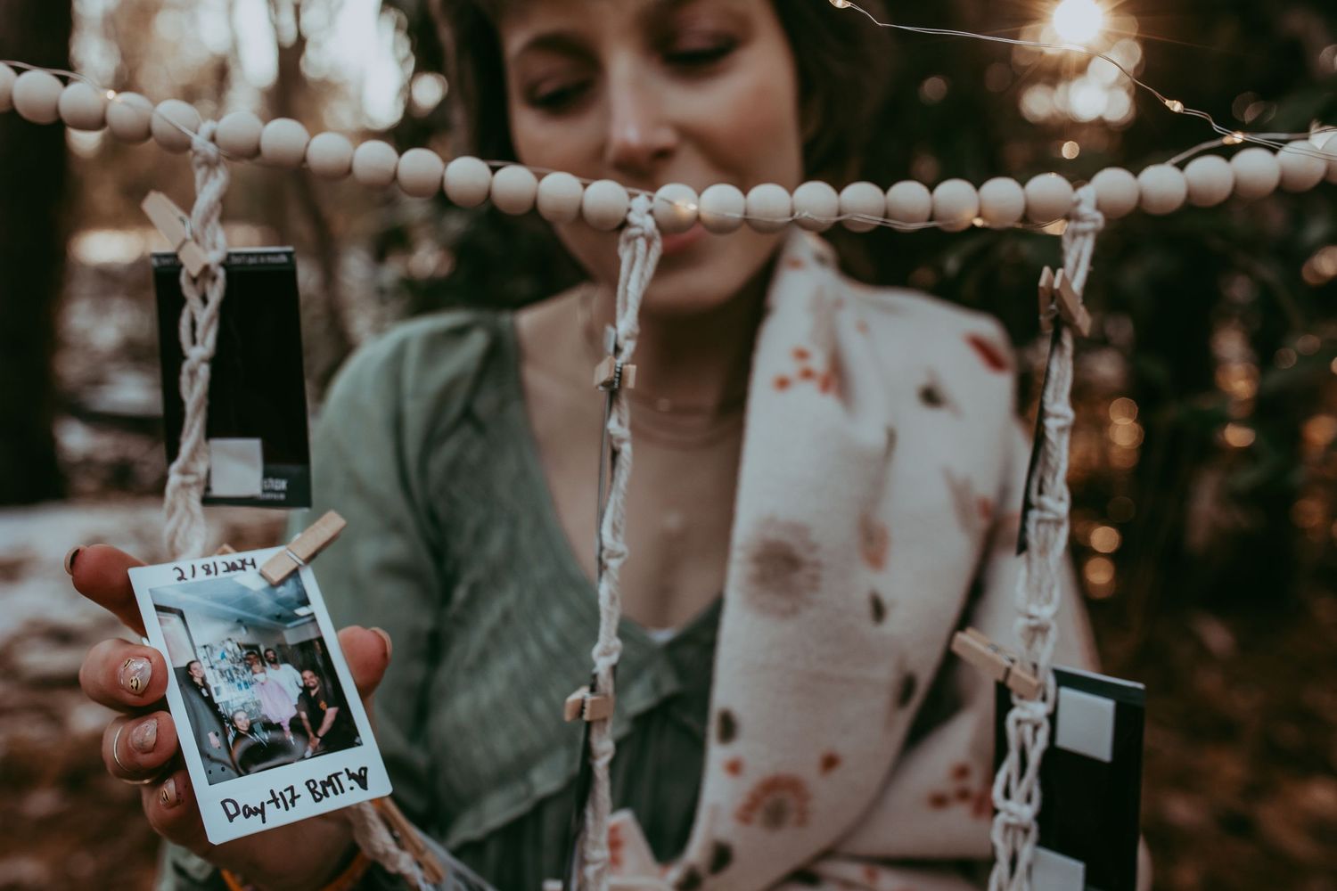 A person hangs vintage polaroid photos on a decorative string with wooden beads outdoors at dusk.